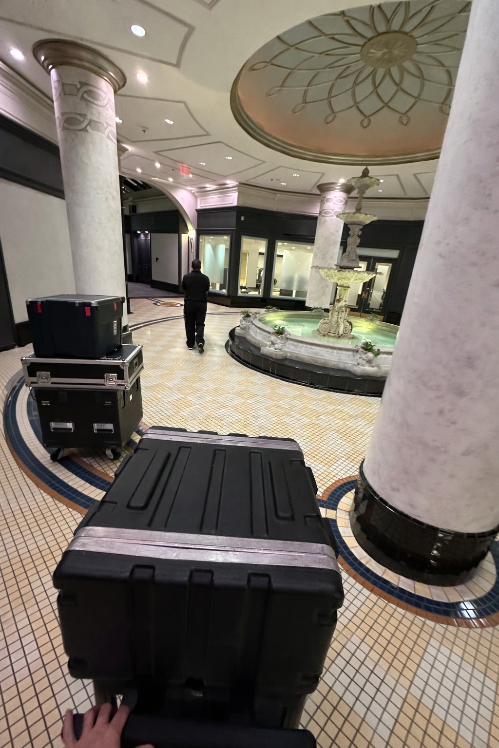 Empty hotel lobby with a fountain, large columns, and decorative ceiling. Equipment is nearby, and a person walks away from the camera.