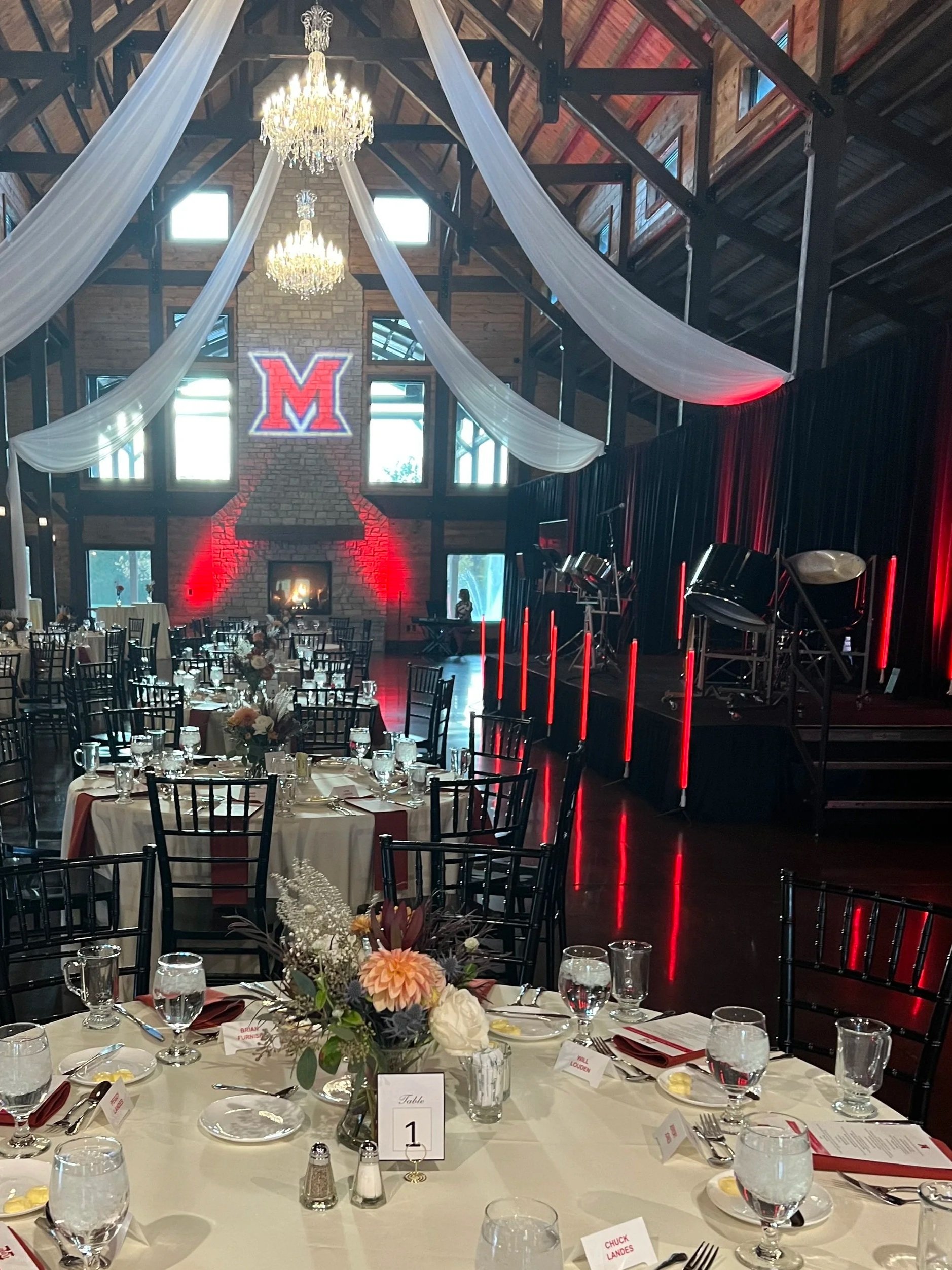 Decorated banquet hall with round tables, floral centerpieces, and place settings, with a stage featuring red lighting, a large 'M' sign, and musical instruments in the background.