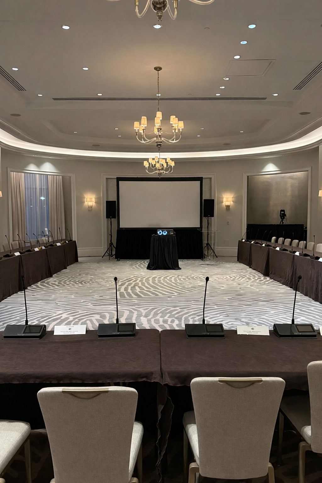 Empty conference room set up for a presentation or meeting with a large screen, microphones on tables, and chairs arranged in a U-shape, inside a well-lit, elegant room with chandeliers and wall sconces.