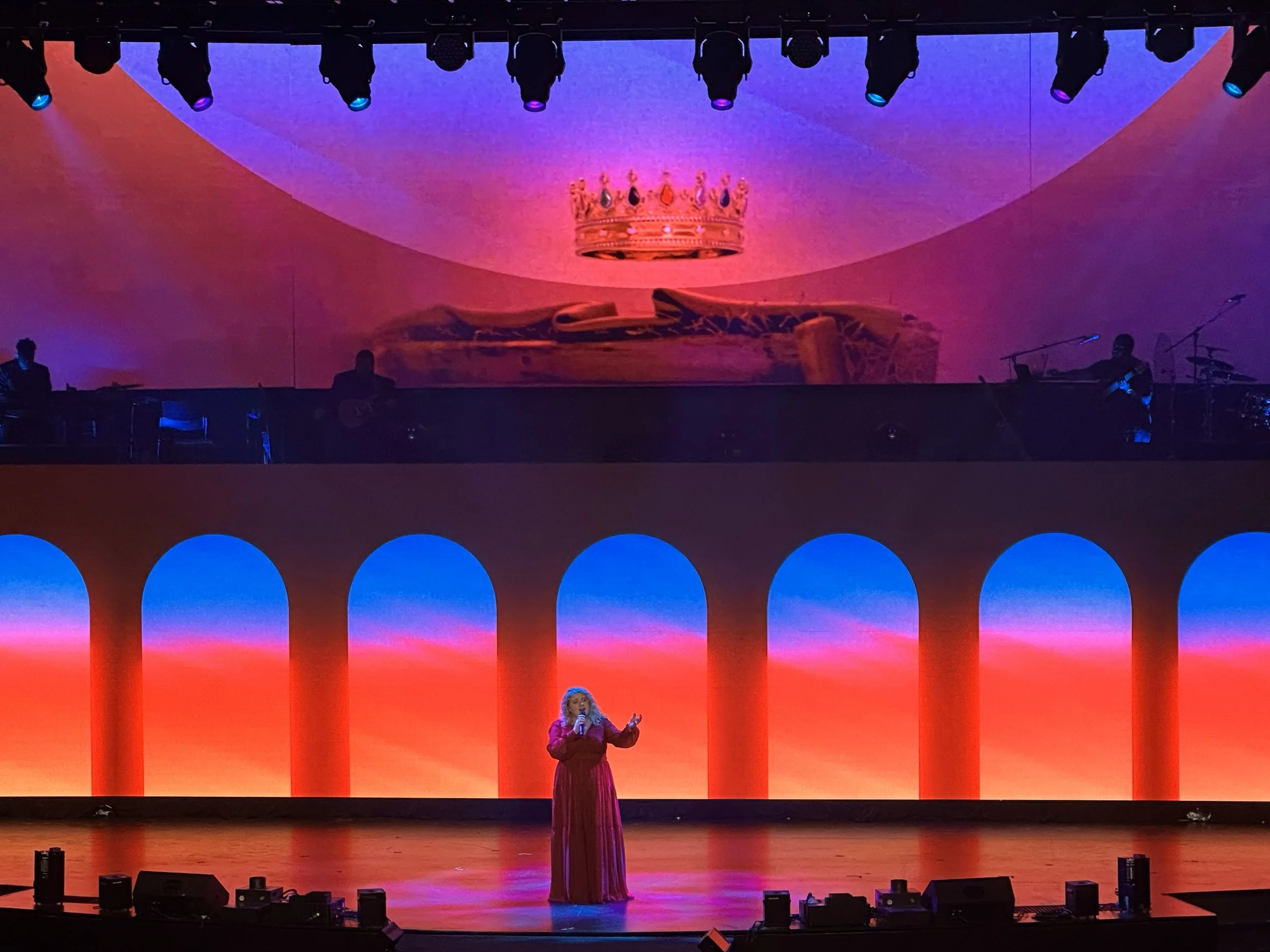 A singer performing on stage in front of a colorful background with arches, and a large screen behind displaying a crown and a sleeping figure. The stage has musicians, and a woman in a red dress is singing in the foreground.