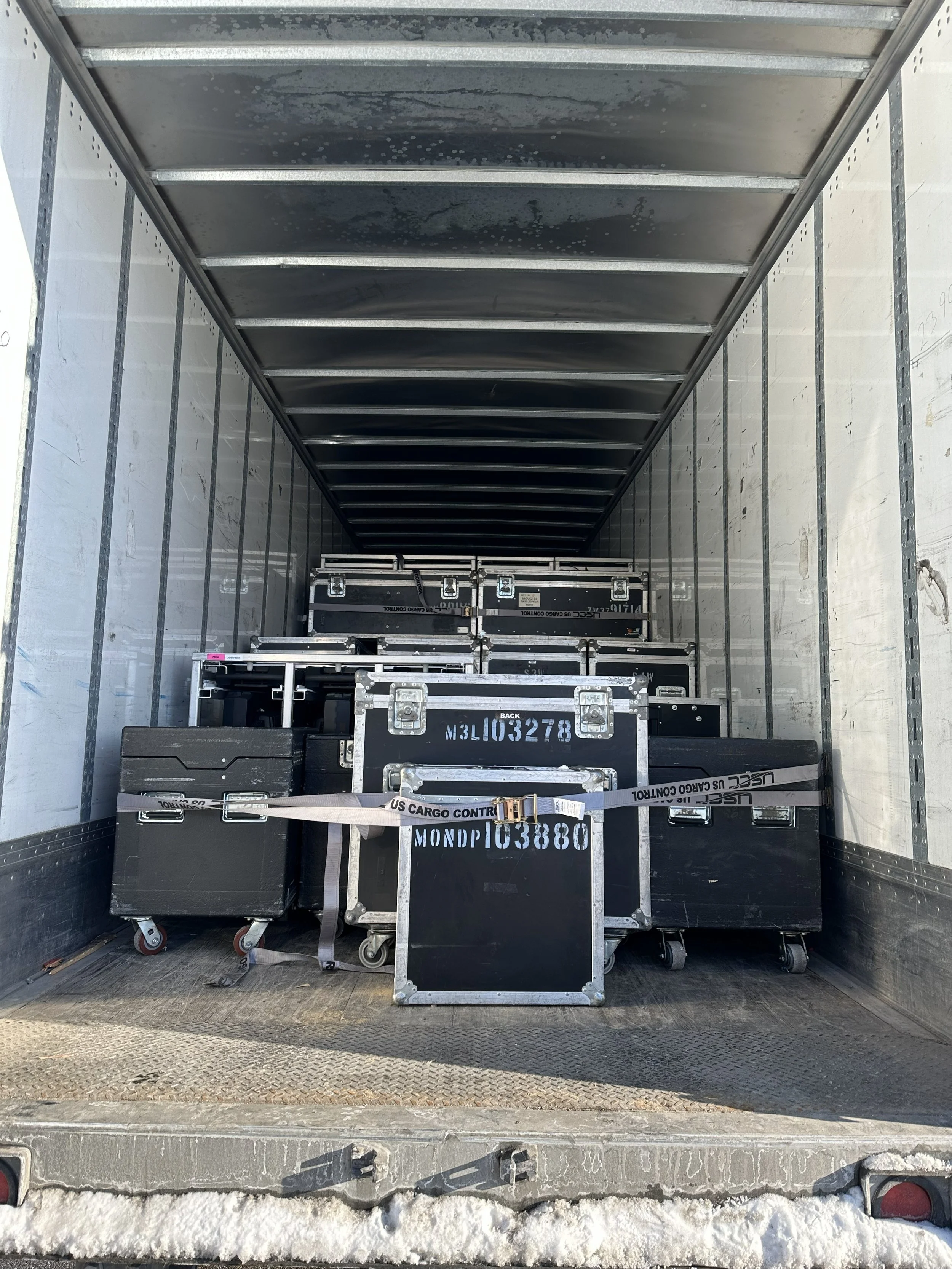 Inside of a truck with black flight cases stacked and secured with cargo straps.