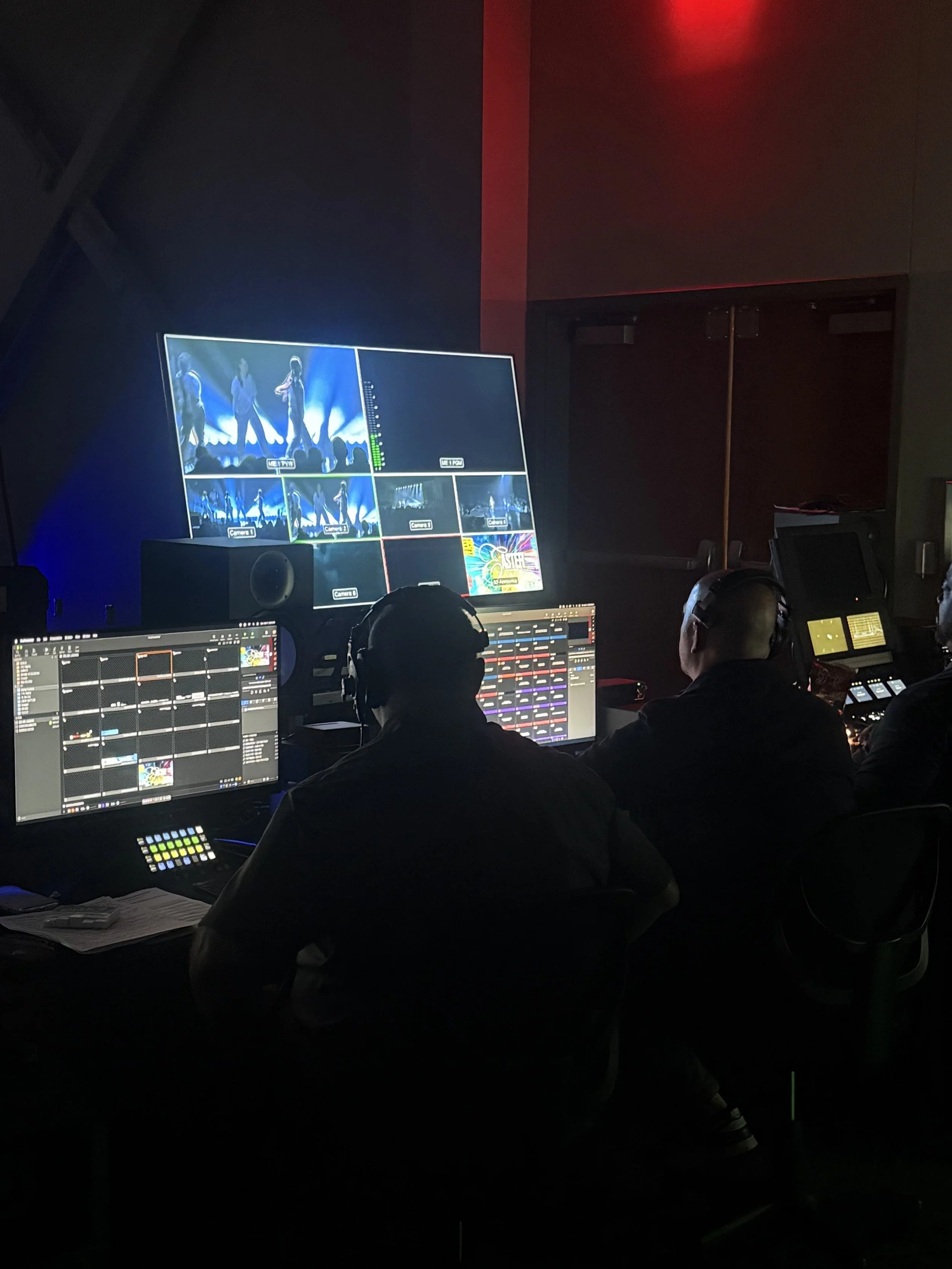 Dark control room with multiple monitors showing a live performance of a band on Stage, crew members work at control panels, with some wearing headphones.