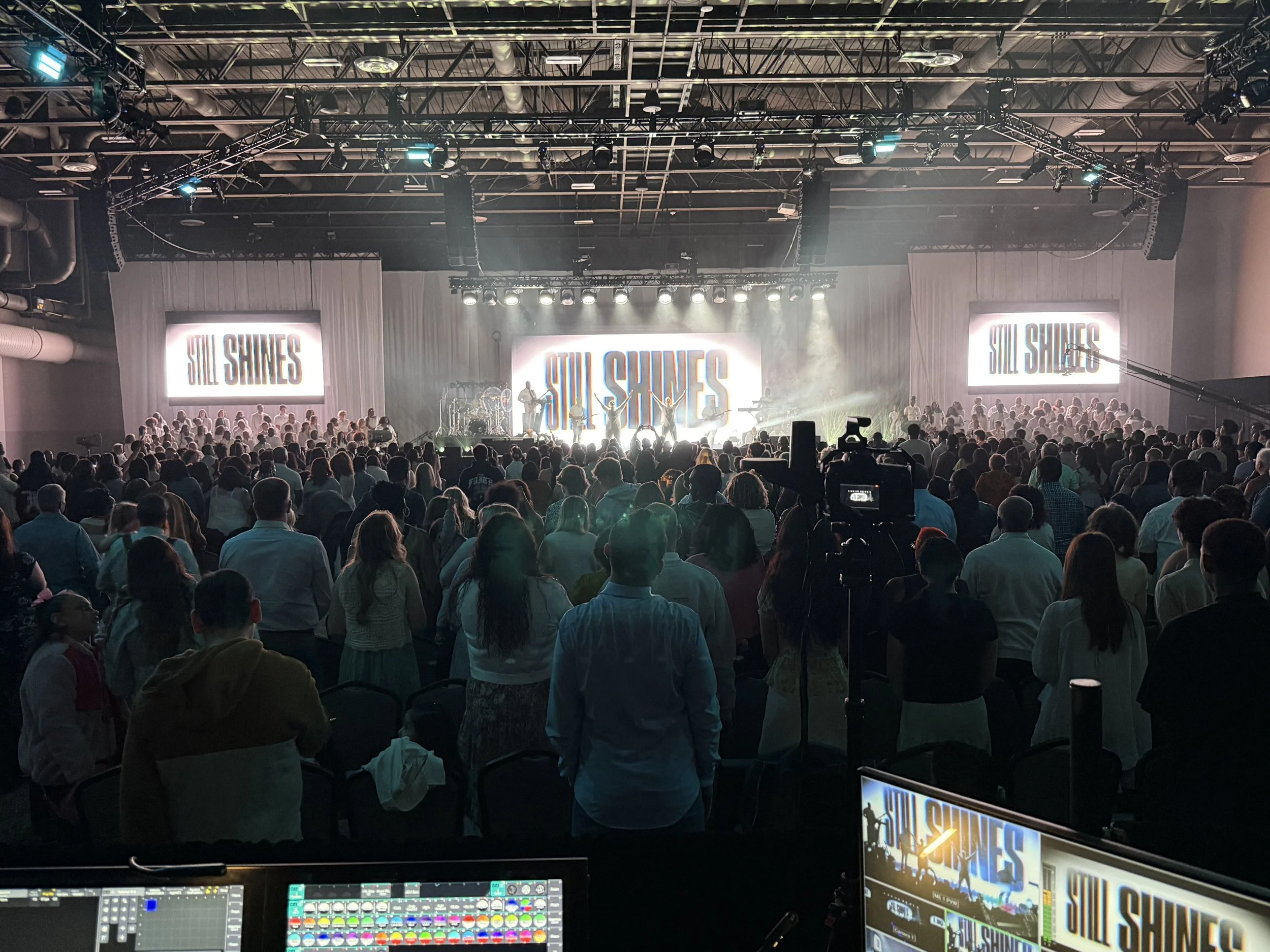A concert stage with a band performing, large screens displaying 'Still Shines' behind them, and a large audience watching. The venue has a high ceiling with stage lighting and sound equipment.