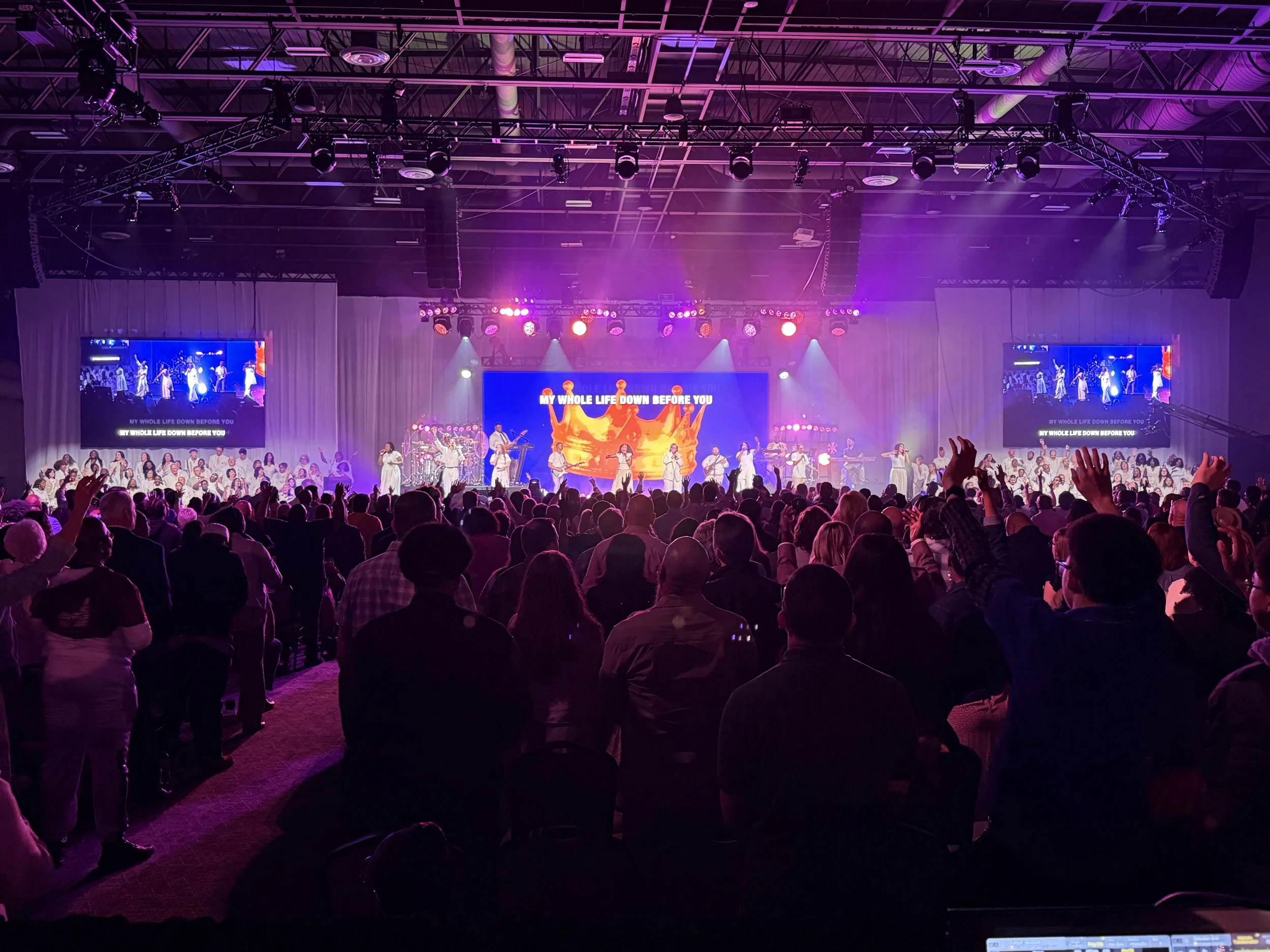 A large audience gathered in a hall watching a stage performance with singers and musicians. The stage has purple and pink lighting, with two large screens displaying the performance and the lyrics, 'MY WHOLE LIFE DOWN BEFORE YOU.'