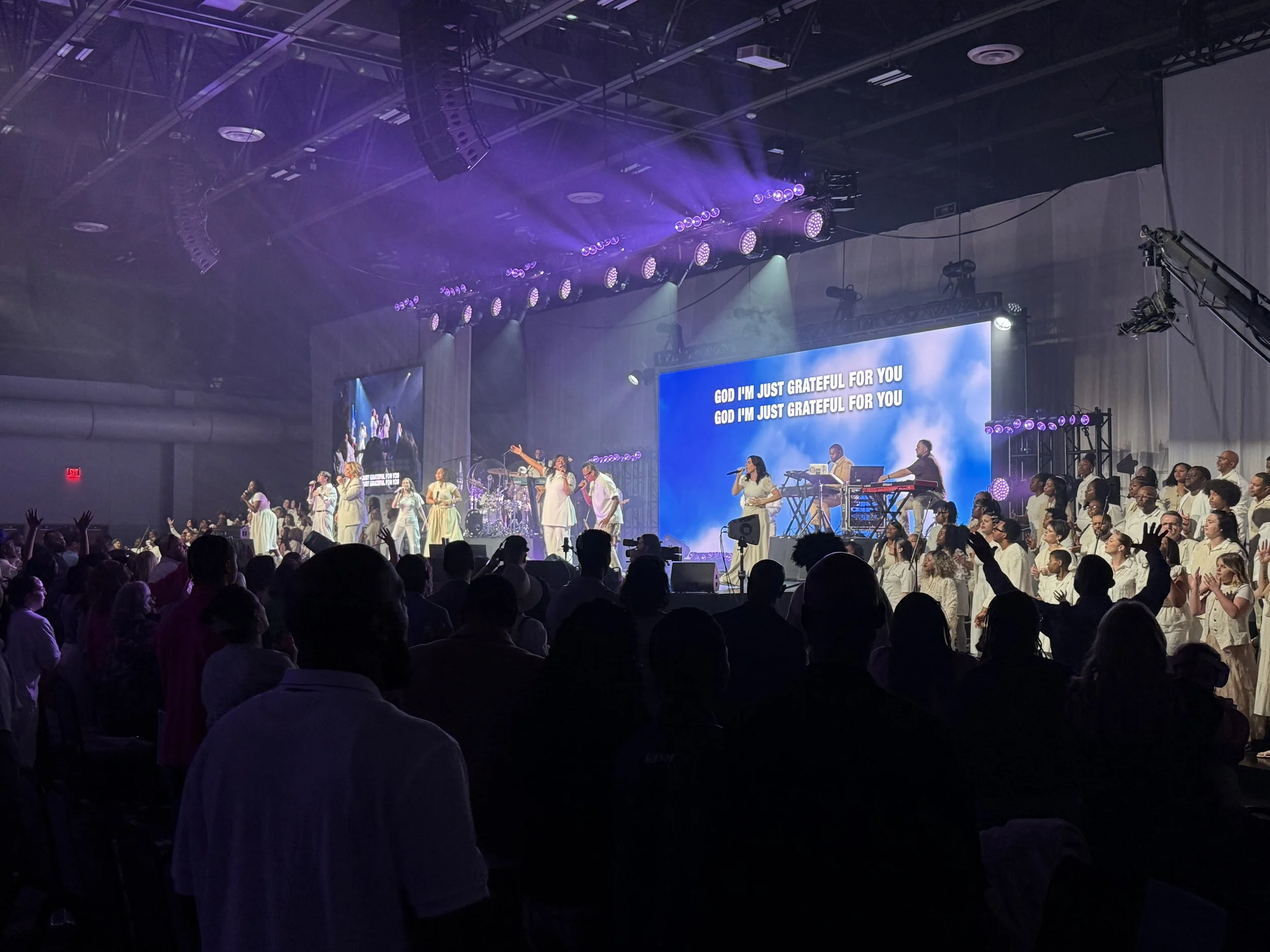 A large indoor concert with many singers and musicians on stage, illuminated by purple and white stage lights, with a crowd of audience members in the foreground, some with raised hands, and a big screen displaying song lyrics in the background.