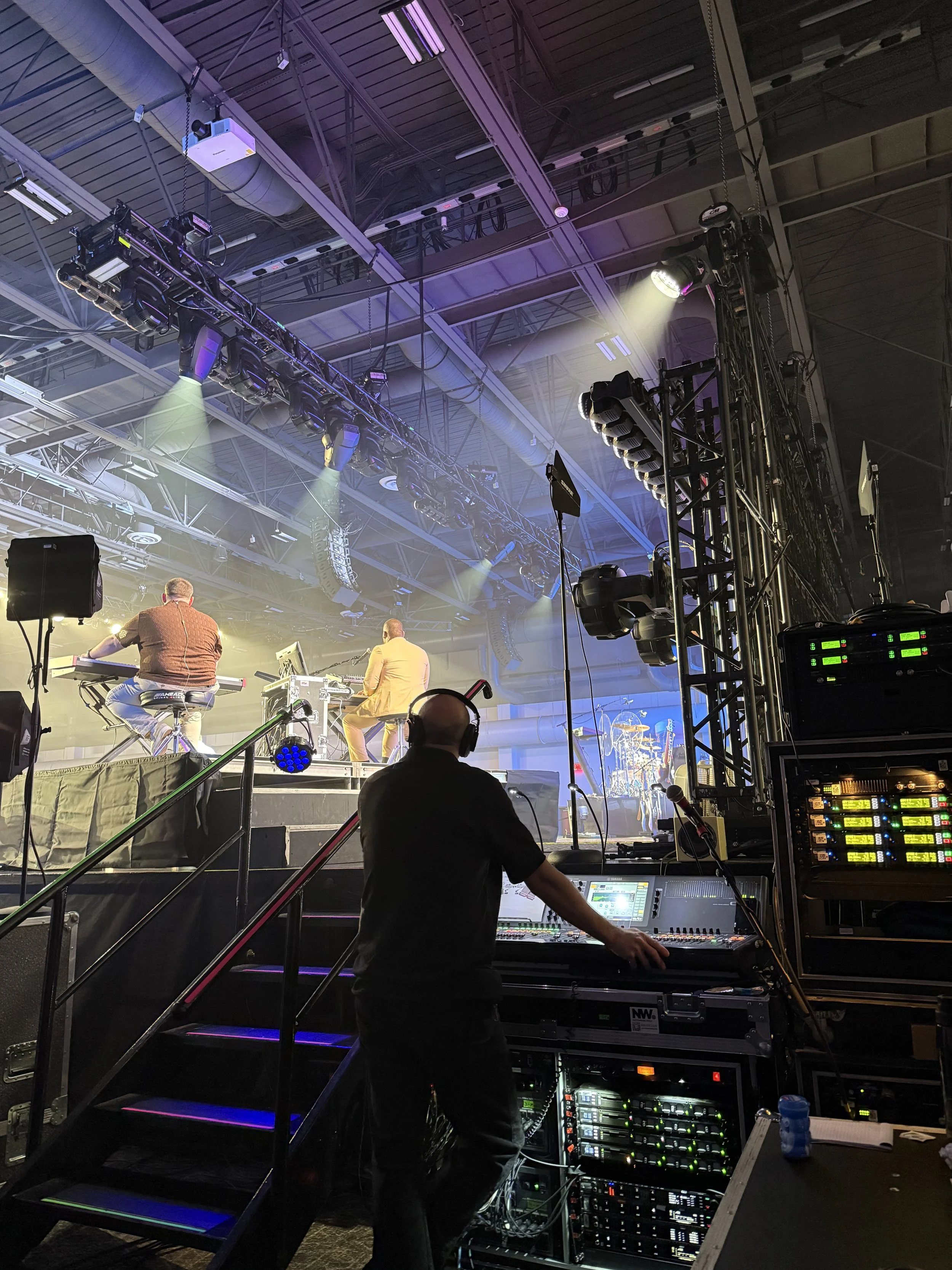 A stage setup with lighting and sound equipment, two musicians playing instruments in the background, and a sound engineer working at a mixing console in the foreground. The ceiling has metallic beams, cables, and mounted lights.