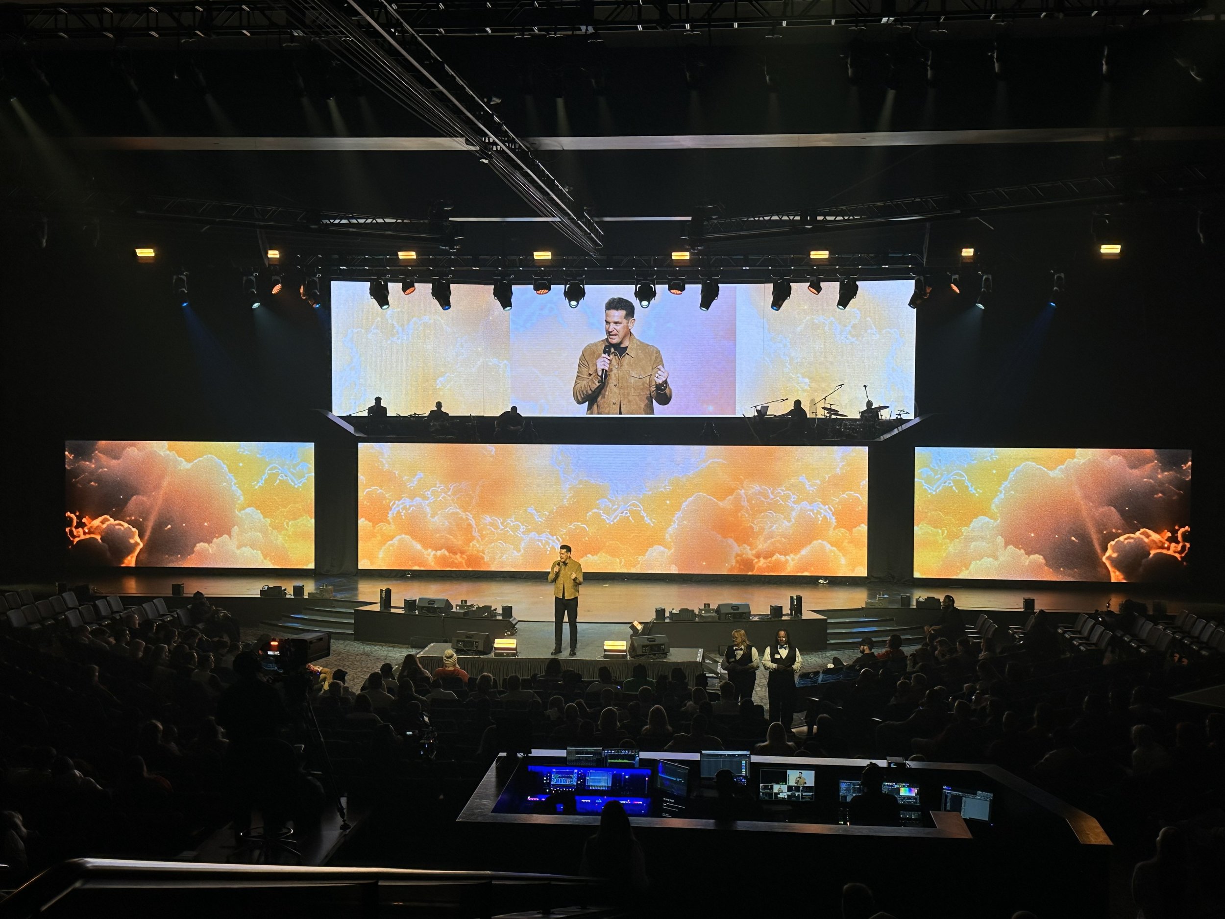 A speaker on stage delivering a presentation at a conference with three large screens displaying an orange and yellow sunset sky with clouds behind him. The audience is seated in front of the stage, and there are event staff and technical equipment v