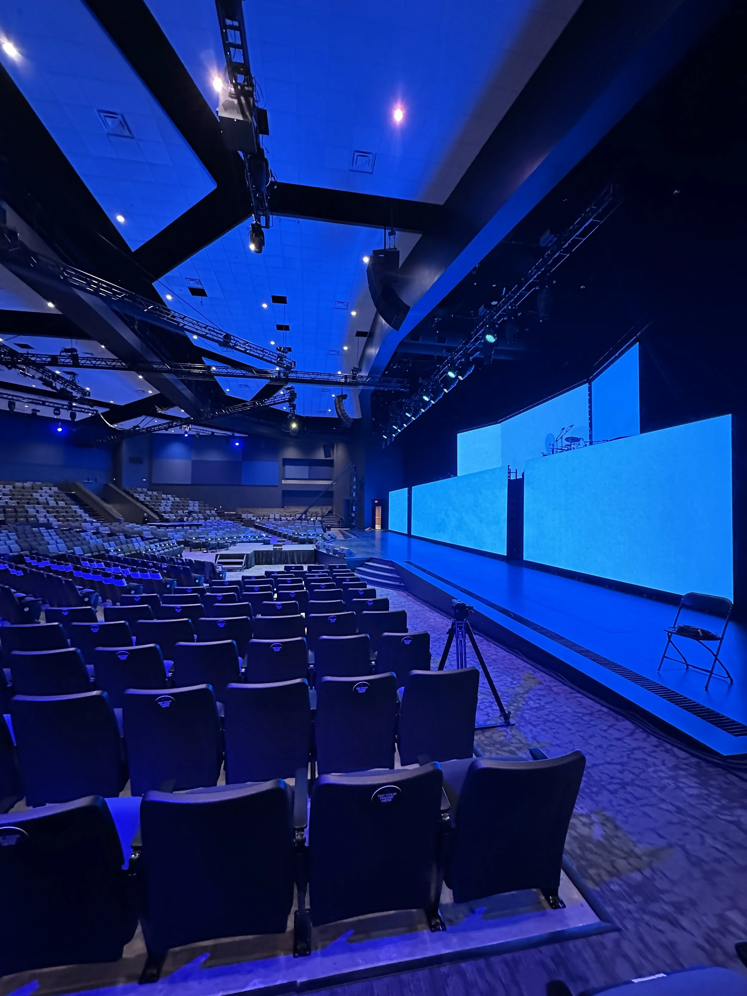 Empty auditorium with rows of black chairs facing a large stage with multiple blue screens and musical instruments, dim blue lighting.
