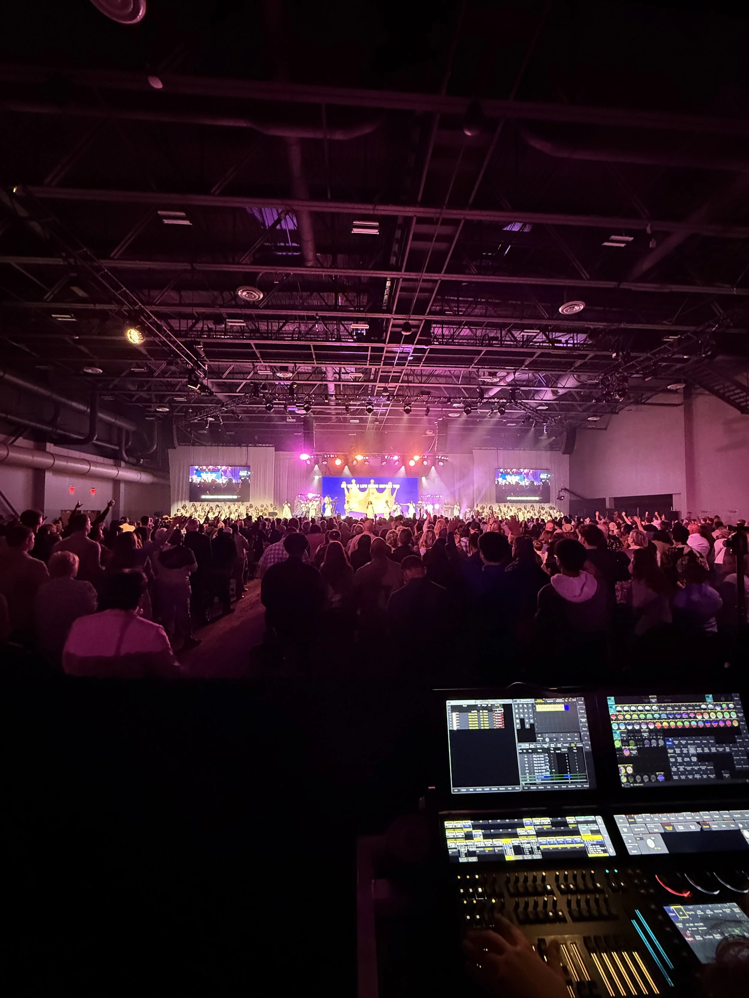 Indoor concert or event venue with a crowd facing a brightly lit stage, visible audio and lighting equipment, and screens displaying visuals.