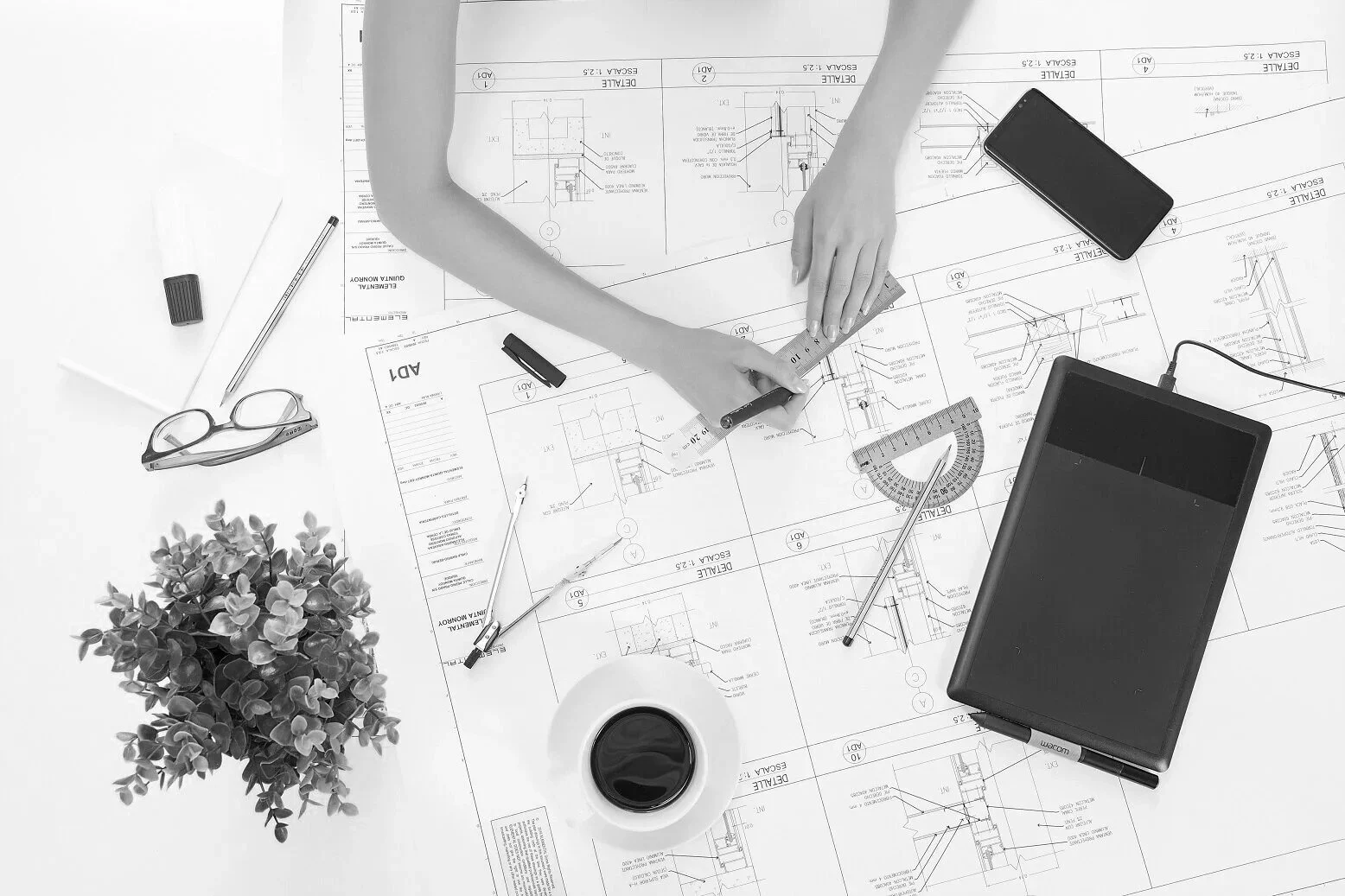 Top view of a desk with architectural blueprints, a person's arms measuring a line, a smartphone, calculator, razors, a pair of glasses, a plant, and a cup of coffee.