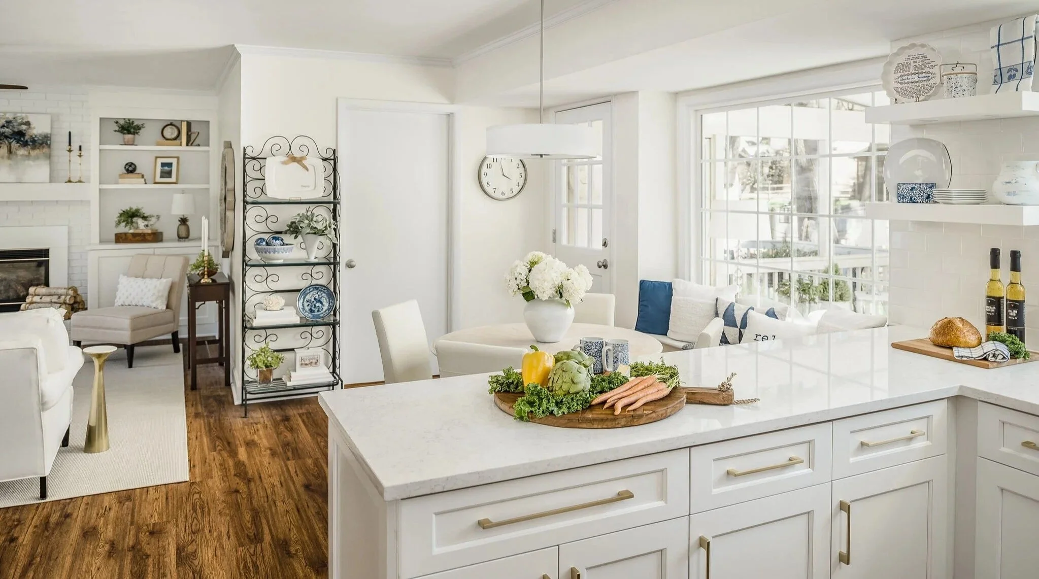 Bright kitchen and dining area with white cabinets, countertops, and a large window with natural light, decorated with fresh vegetables, flowers, and home decor.