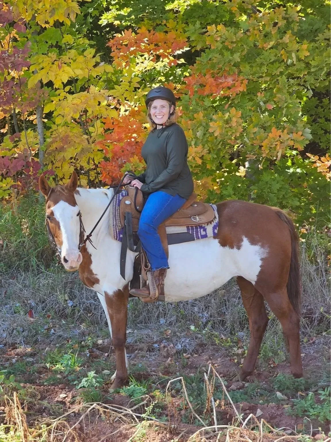 A woman with a helmet smiling while sitting on a brown and white horse, with colorful autumn leaves in the background.