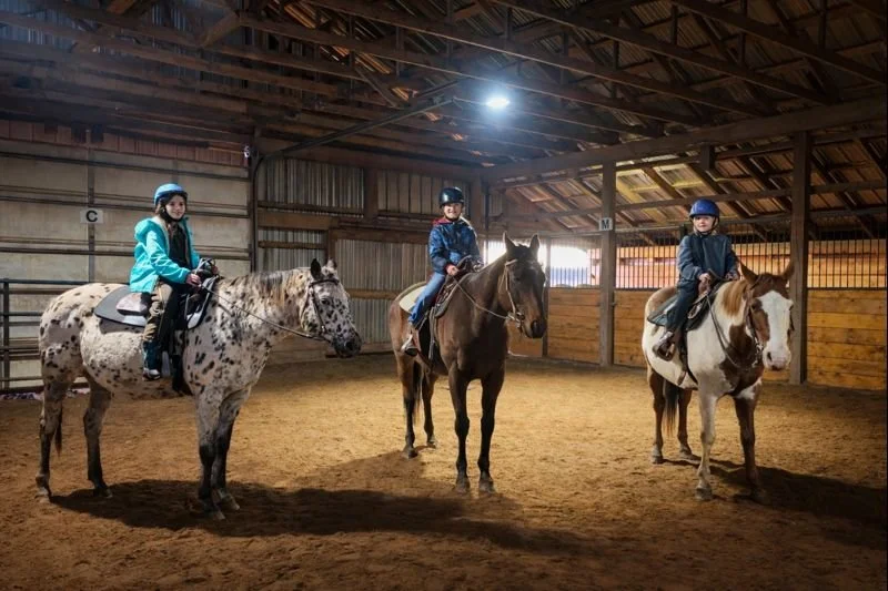 Three children riding horses inside an indoor riding arena.