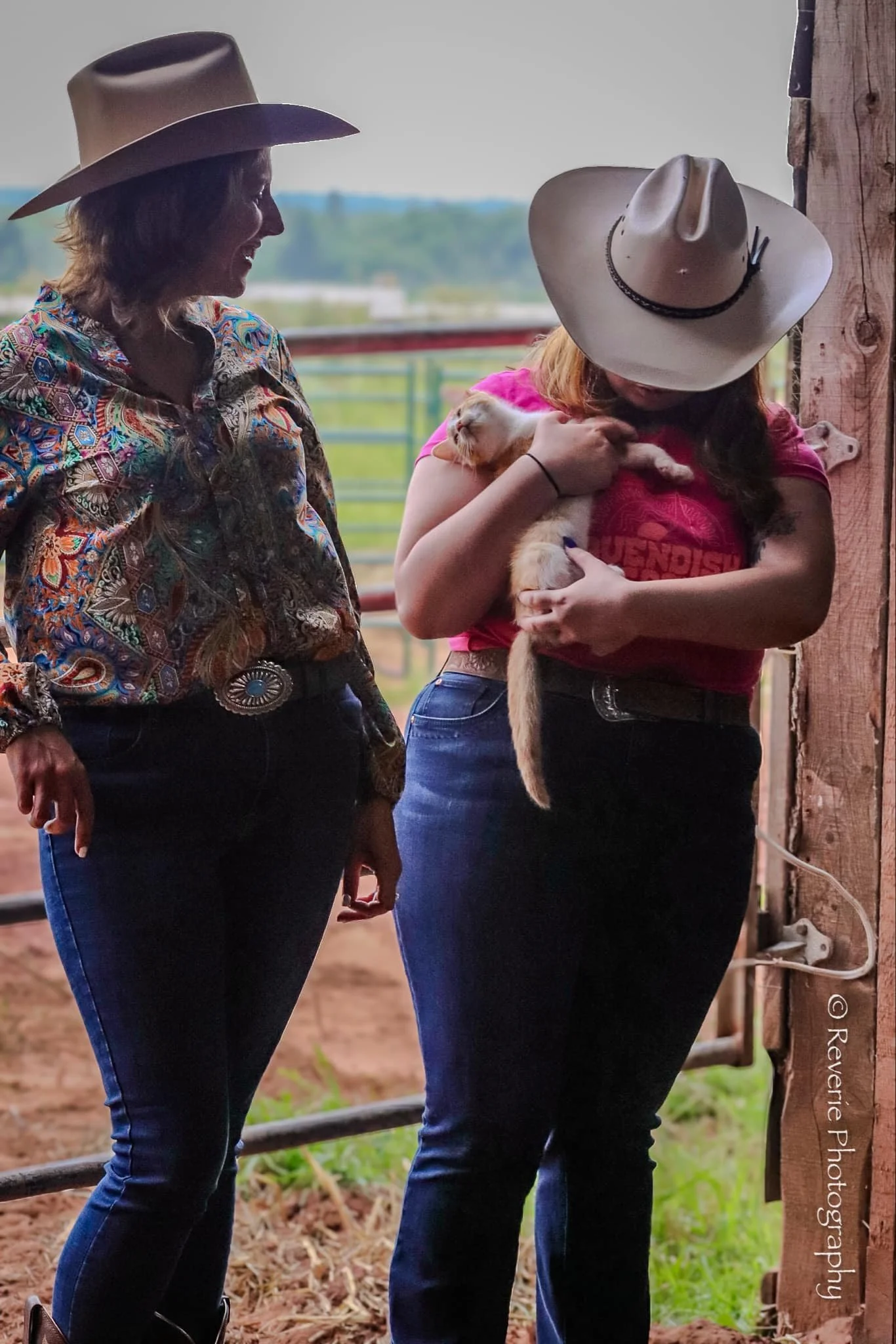 Two women wearing cowboy hats, one with a colorful shirt and the other with a red shirt, standing indoors near a wooden post. The woman in the red shirt is holding a small kitten.