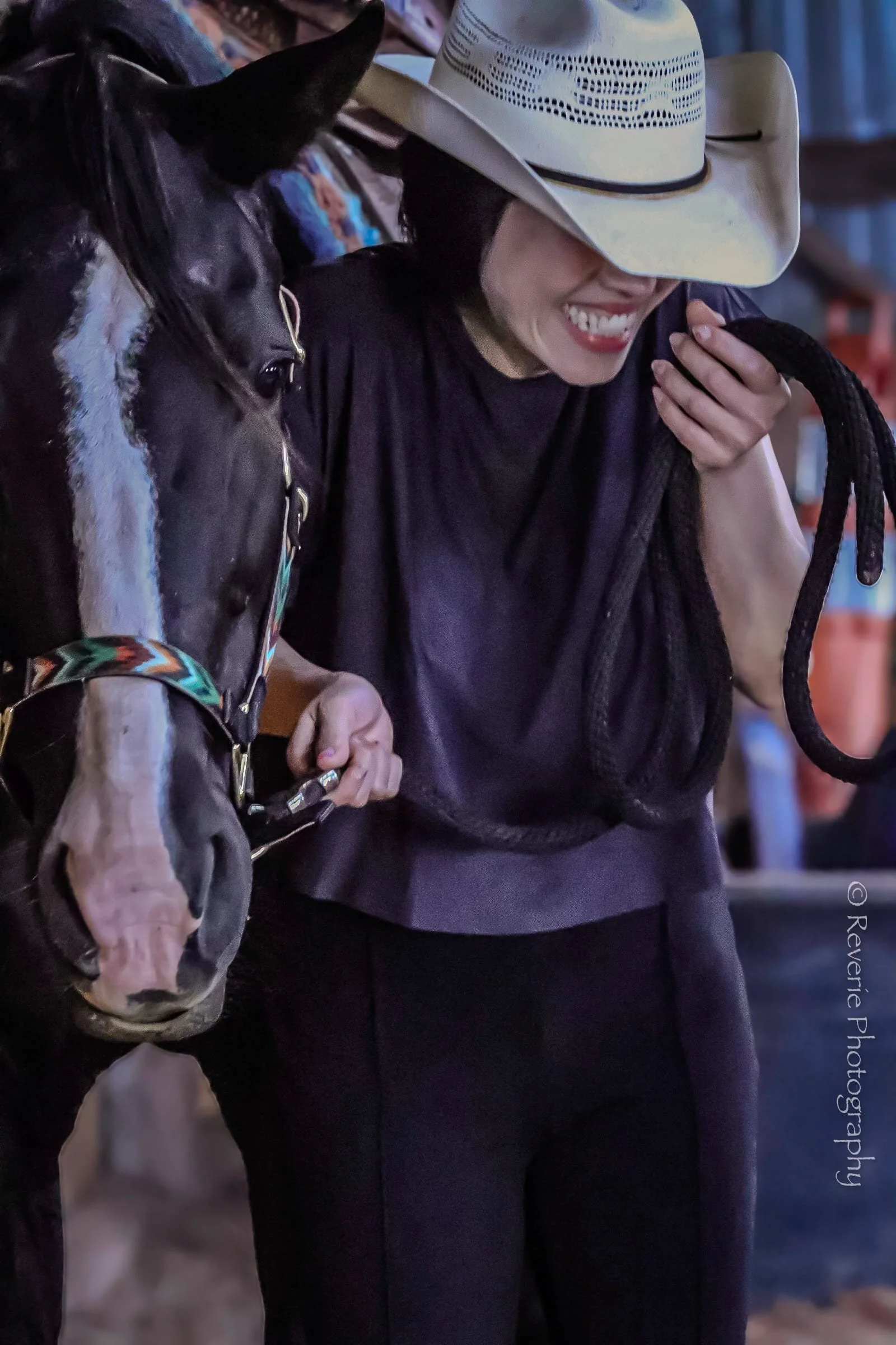 A woman wearing a beige cowboy hat, black shirt, and black pants smiling and looking down while holding a black horse's bridle.