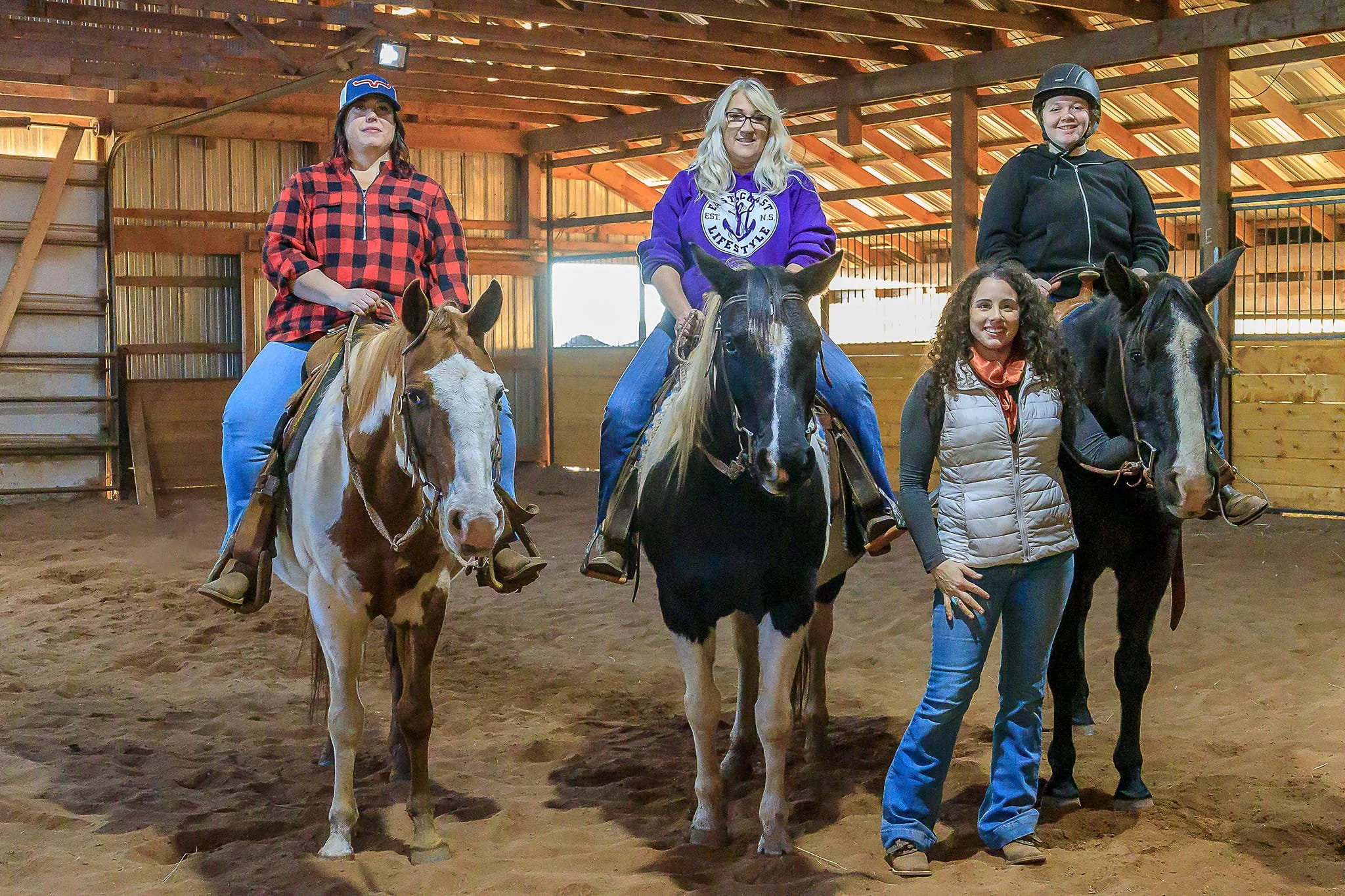 Three women riding horses and one woman standing beside a horse in an indoor riding arena.