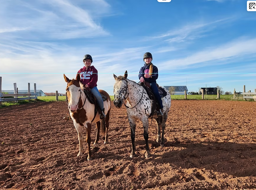 Two children riding horses on a dirt field under a blue sky, with barns and fencing in the background.