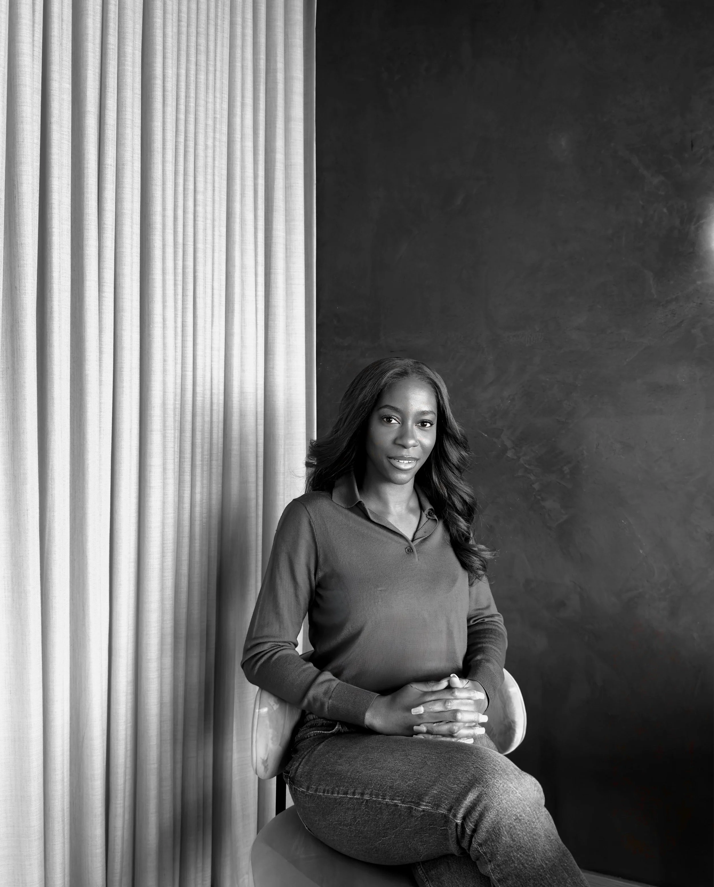 A woman sitting on a chair against a textured dark wall and light curtain, smiling with her hands clasped.