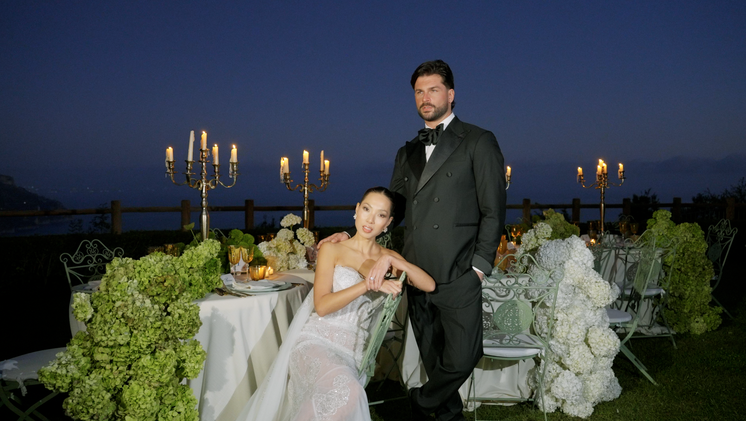 A couple dressed in formal wedding attire at an outdoor evening wedding reception. The woman is sitting in a chair wearing a white wedding gown, and the man is standing beside her in a black tuxedo. The table is decorated with white and green floral arrangements, gold and white candles, and tableware.