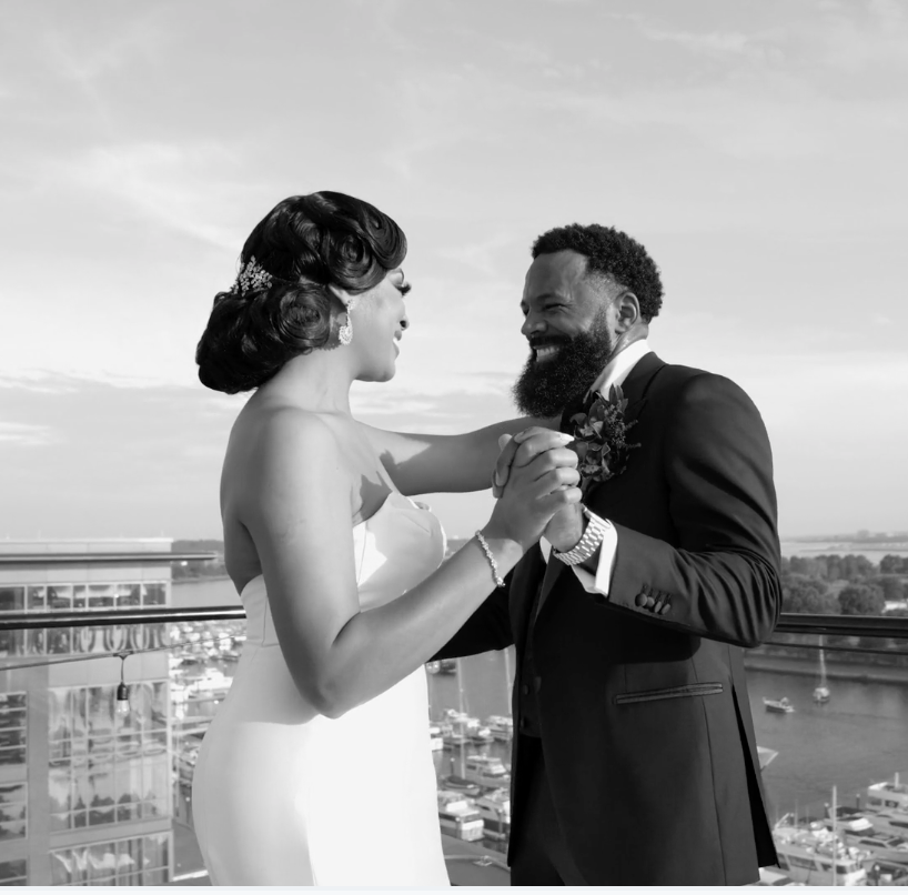 A bride and groom dancing and smiling on an outdoor balcony with a city view and water in the background, in black and white.