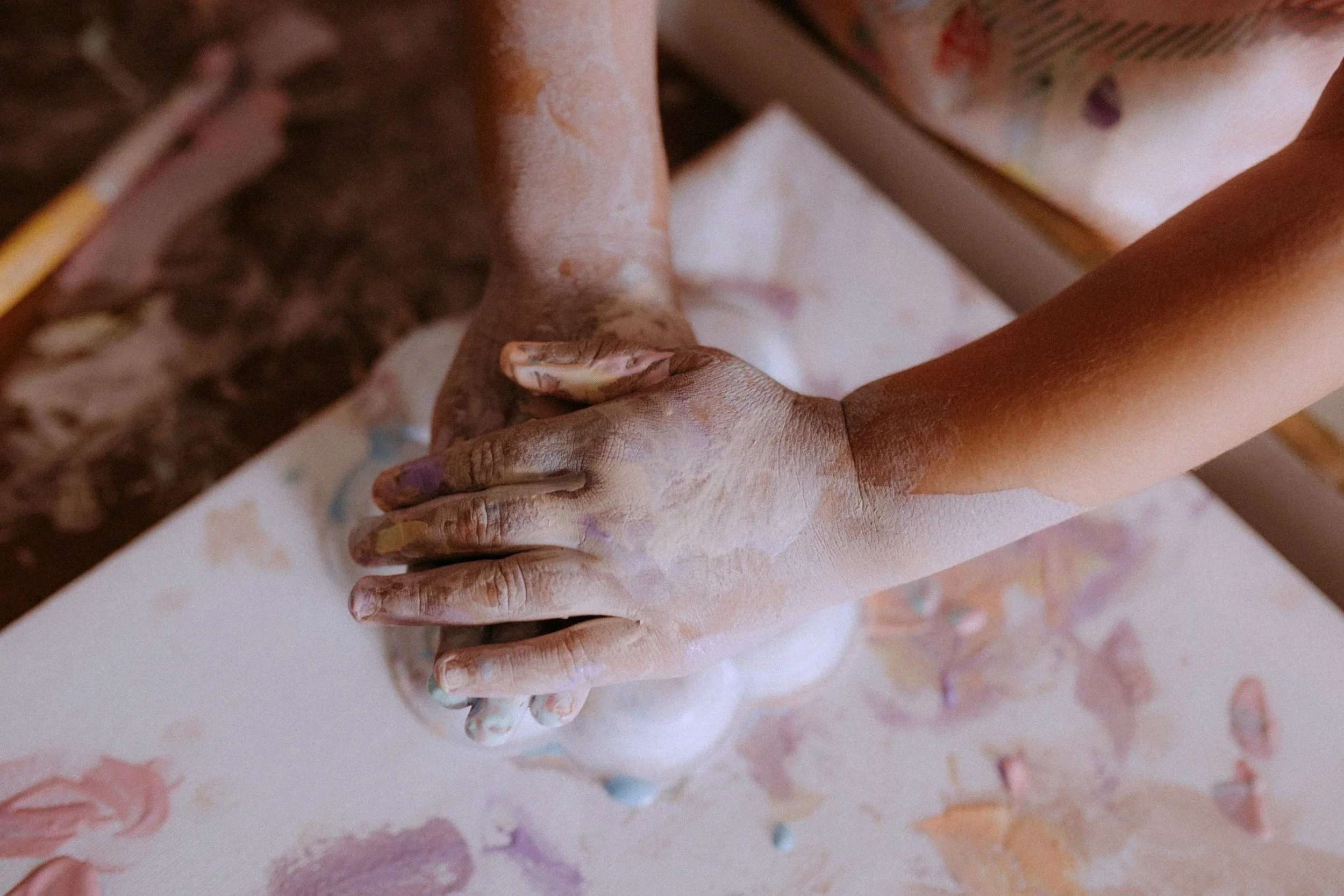 Two hands, one young and one older, are pressing together with fingers intertwined, covered in colorful paint, on a white surface with paint splatters.