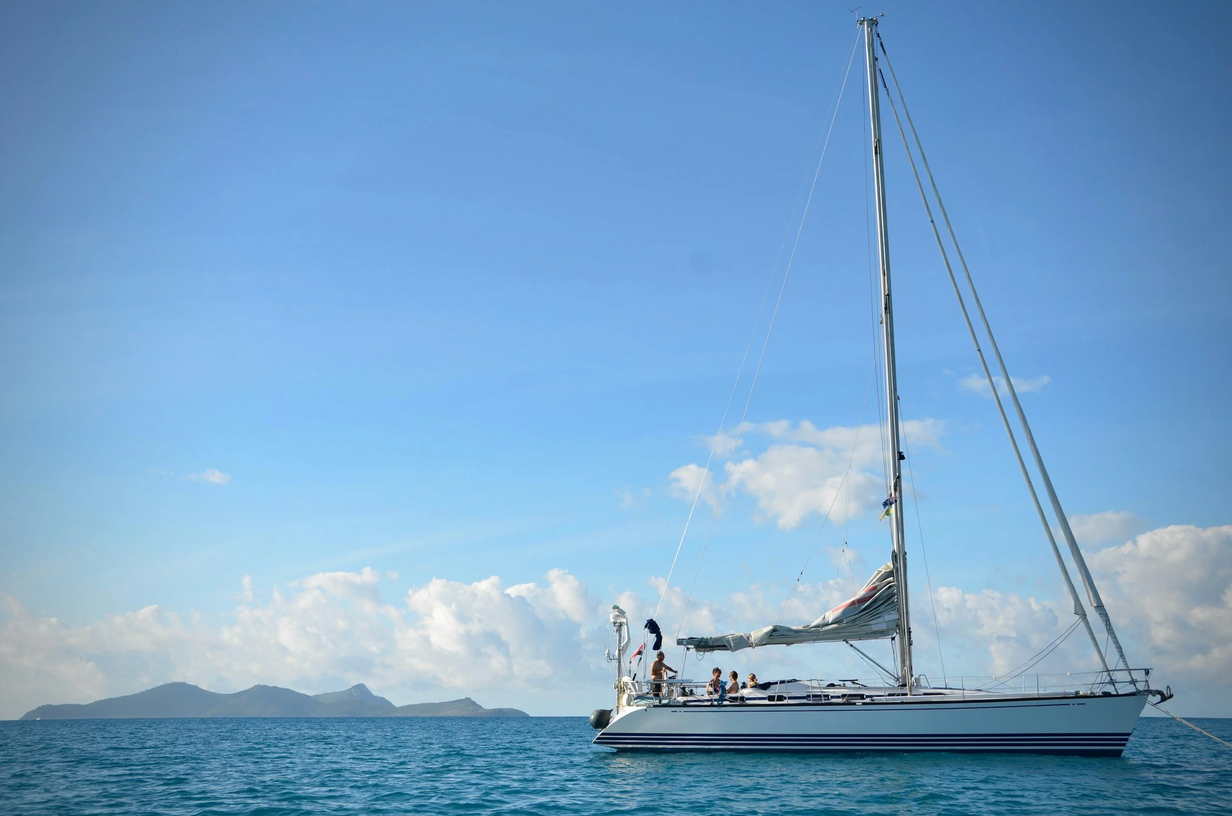 People on a sailboat in calm ocean water with an island and mountains in the distance under a partly cloudy sky.