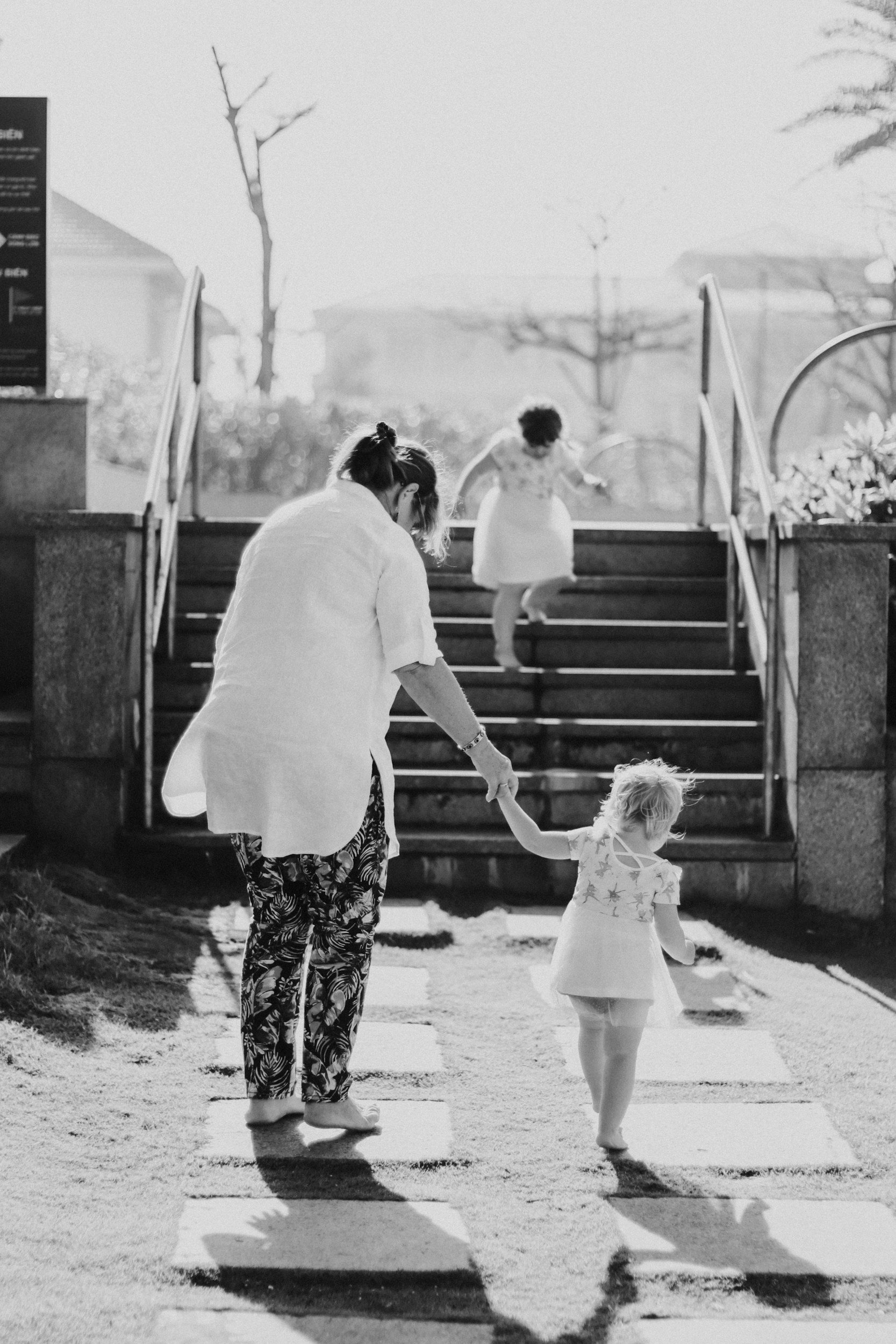 A black and white photo of an adult holding hands with a small child walking on a stepping stone path. Two other children are going up stairs in the background. The scene is outdoors with sun and trees.