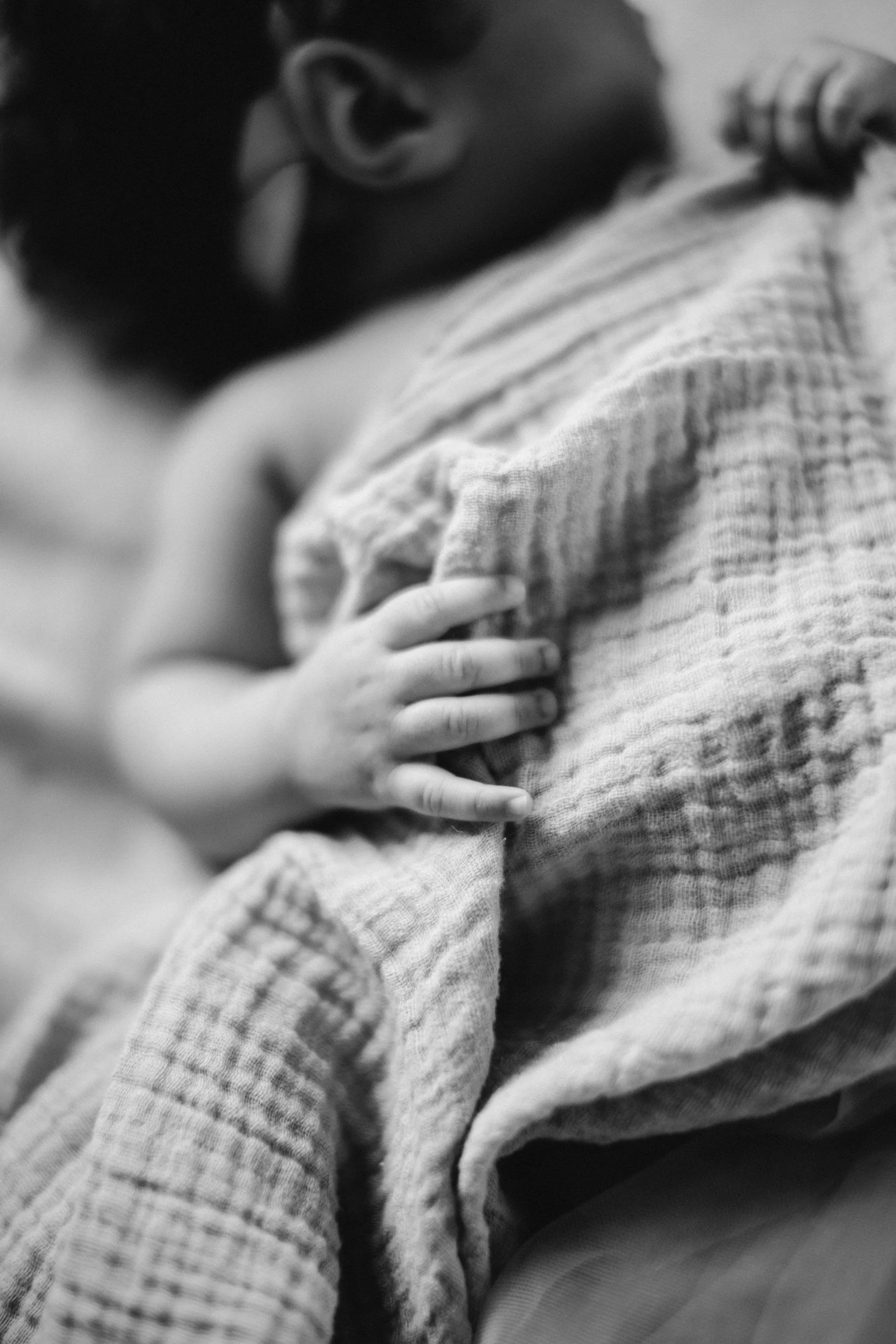 A close-up black and white photo of a small child's hand resting on an adult's shoulder, with the child's face out of focus in the background.