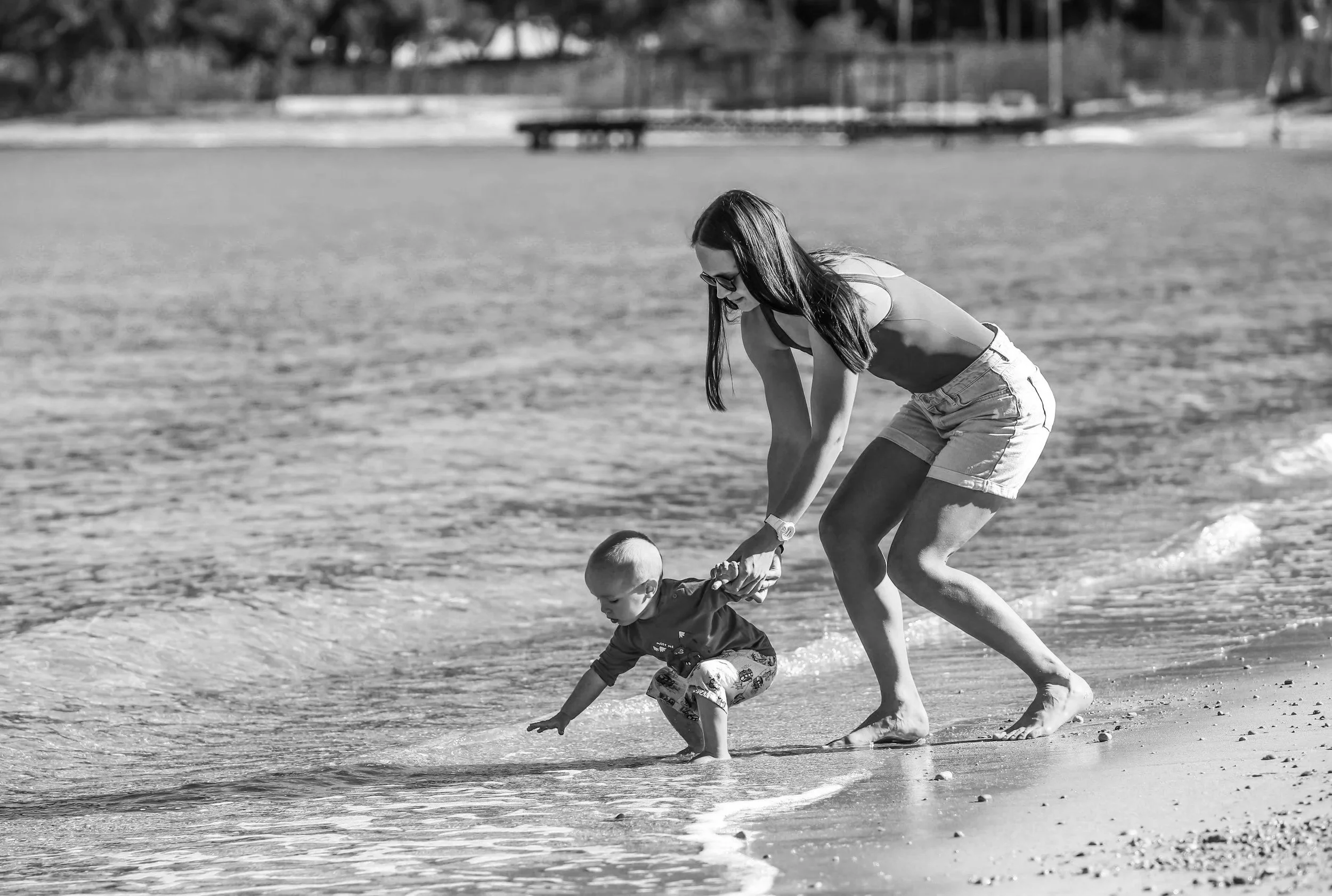 A woman and a young boy playing together at the beach, with the woman helping the boy as he reaches into the water.
