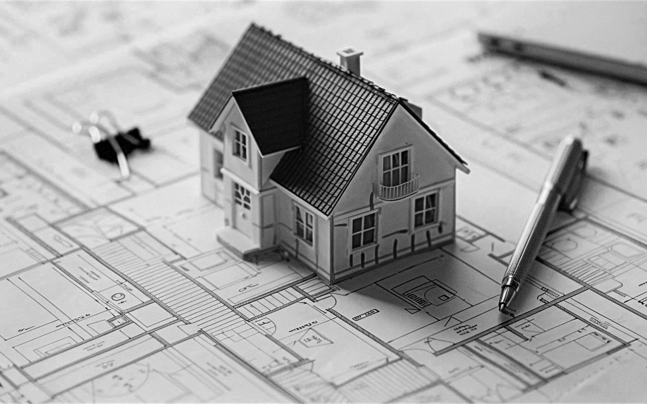 A small model house placed on an architectural blueprint, with a pen resting on the blueprint and a binder clip in the background.