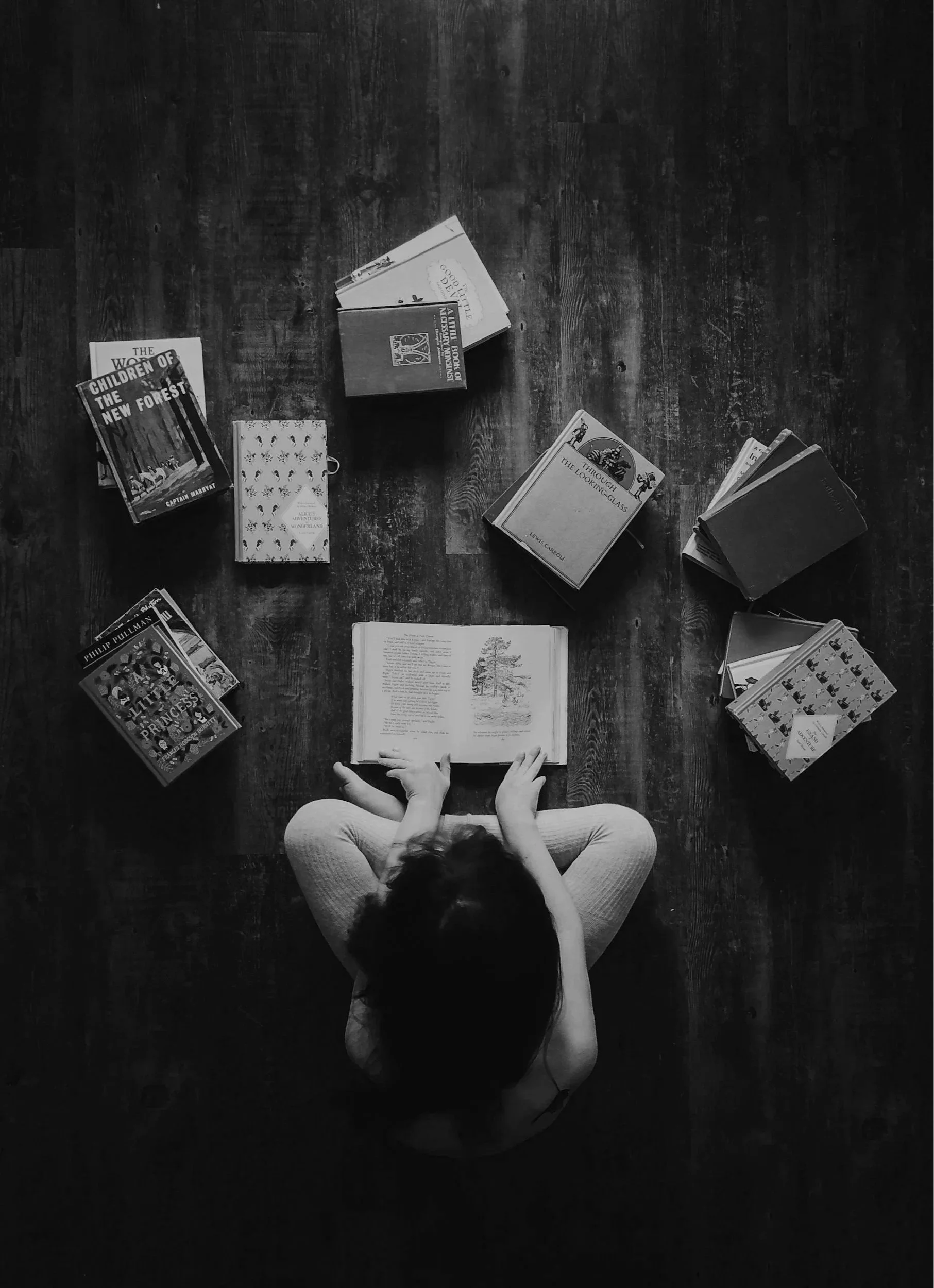 A person sitting on a wooden floor reading a book, surrounded by stacks of books in a circle around them.