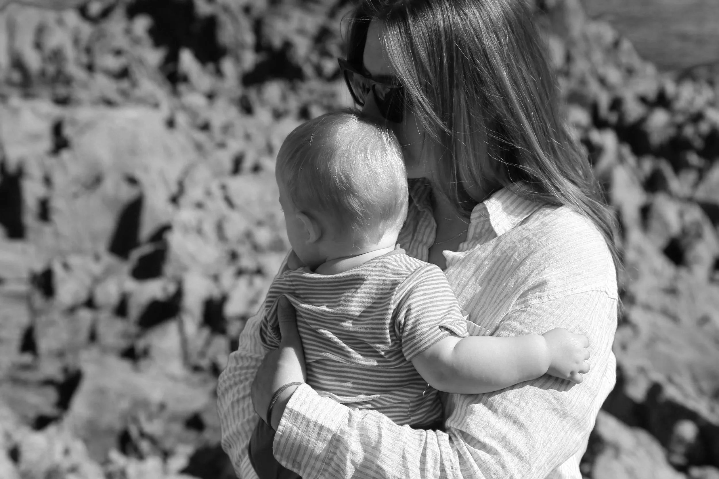 A woman holding a baby close, both facing each other, outdoors with rocks in the background, in black and white.