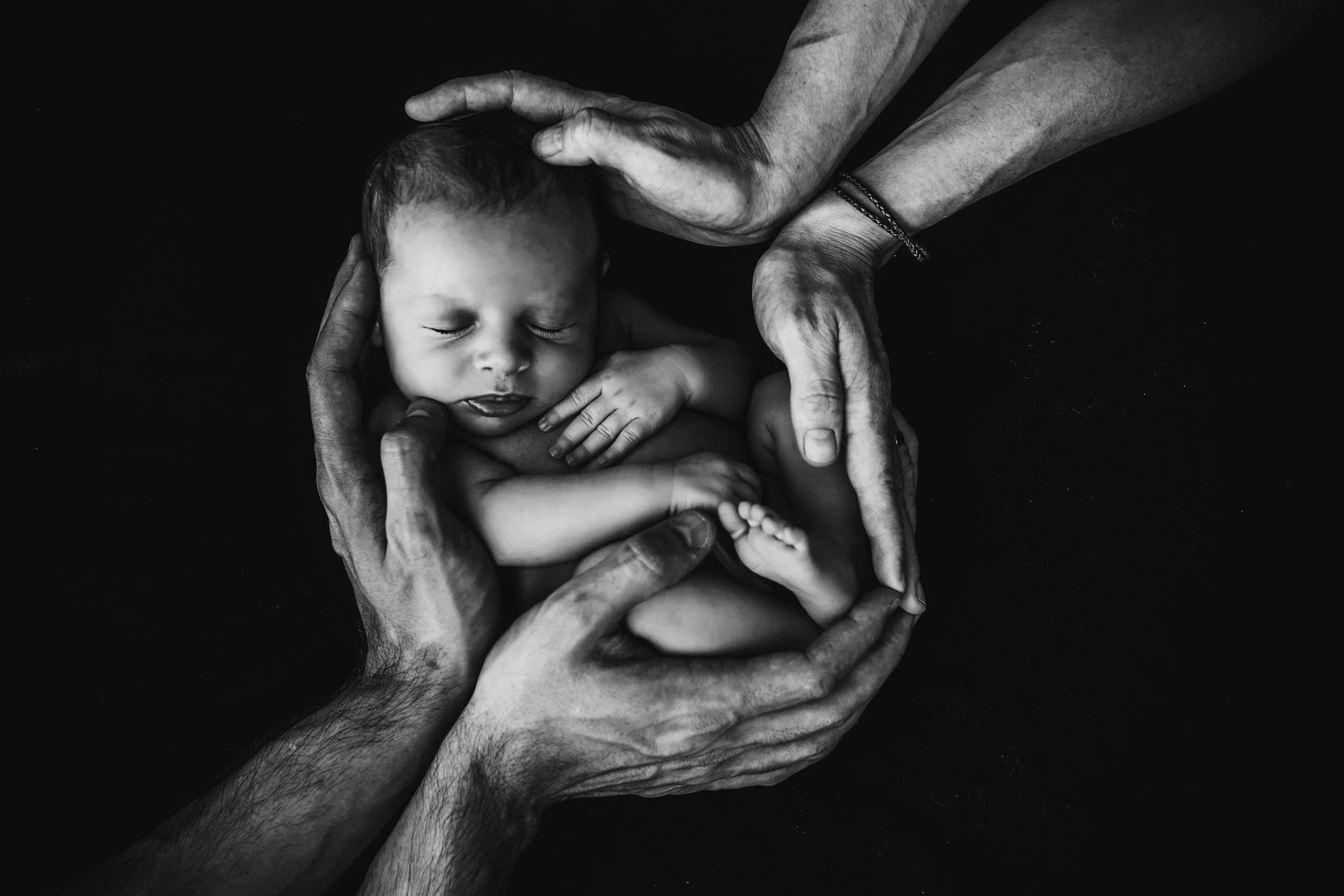 Black and white photo of an infant sleeping, surrounded by the hands of multiple adults gently cradling the baby against a dark background.