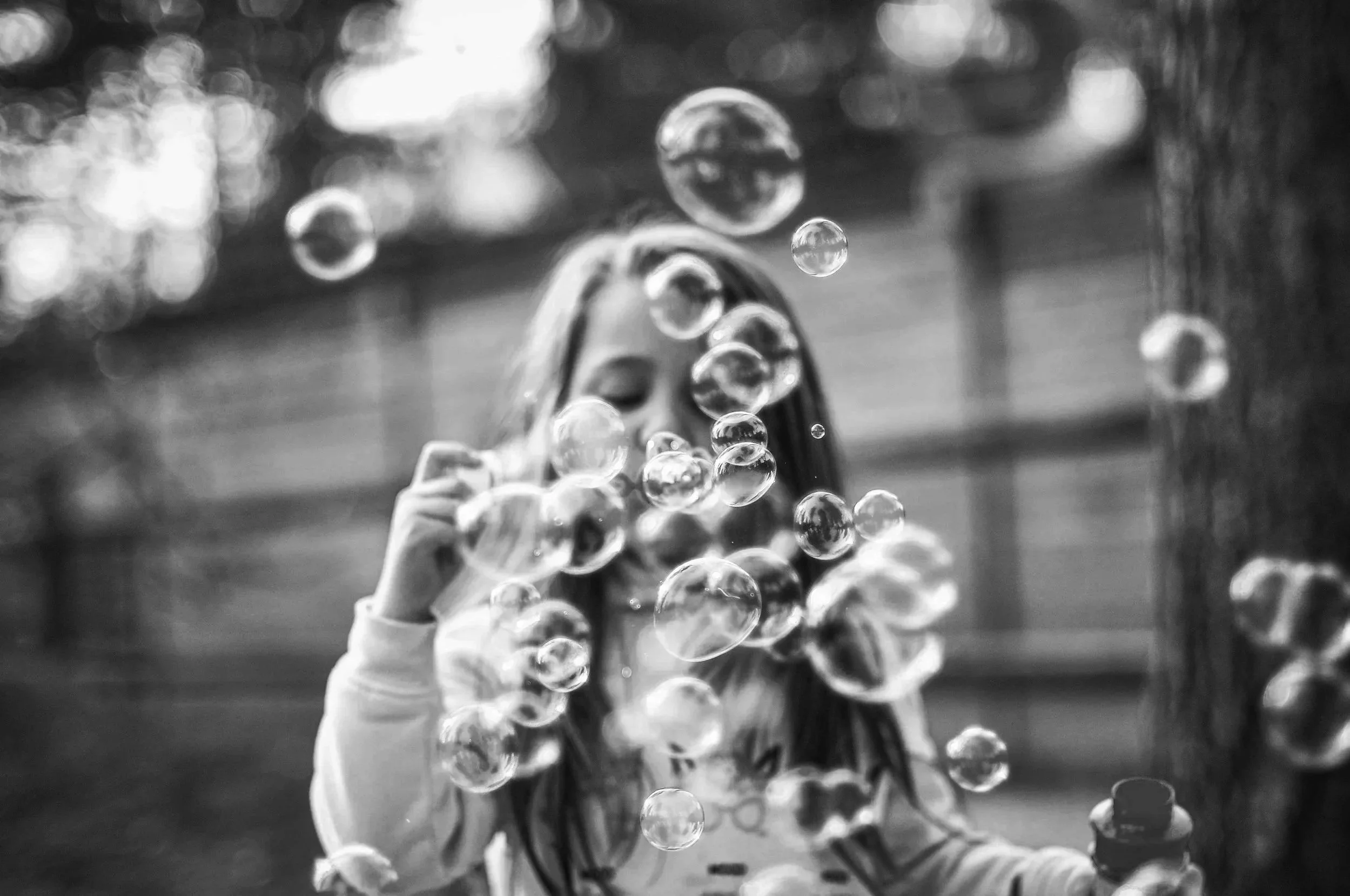 A girl with long hair is blowing bubbles outdoors, with a blurred background.