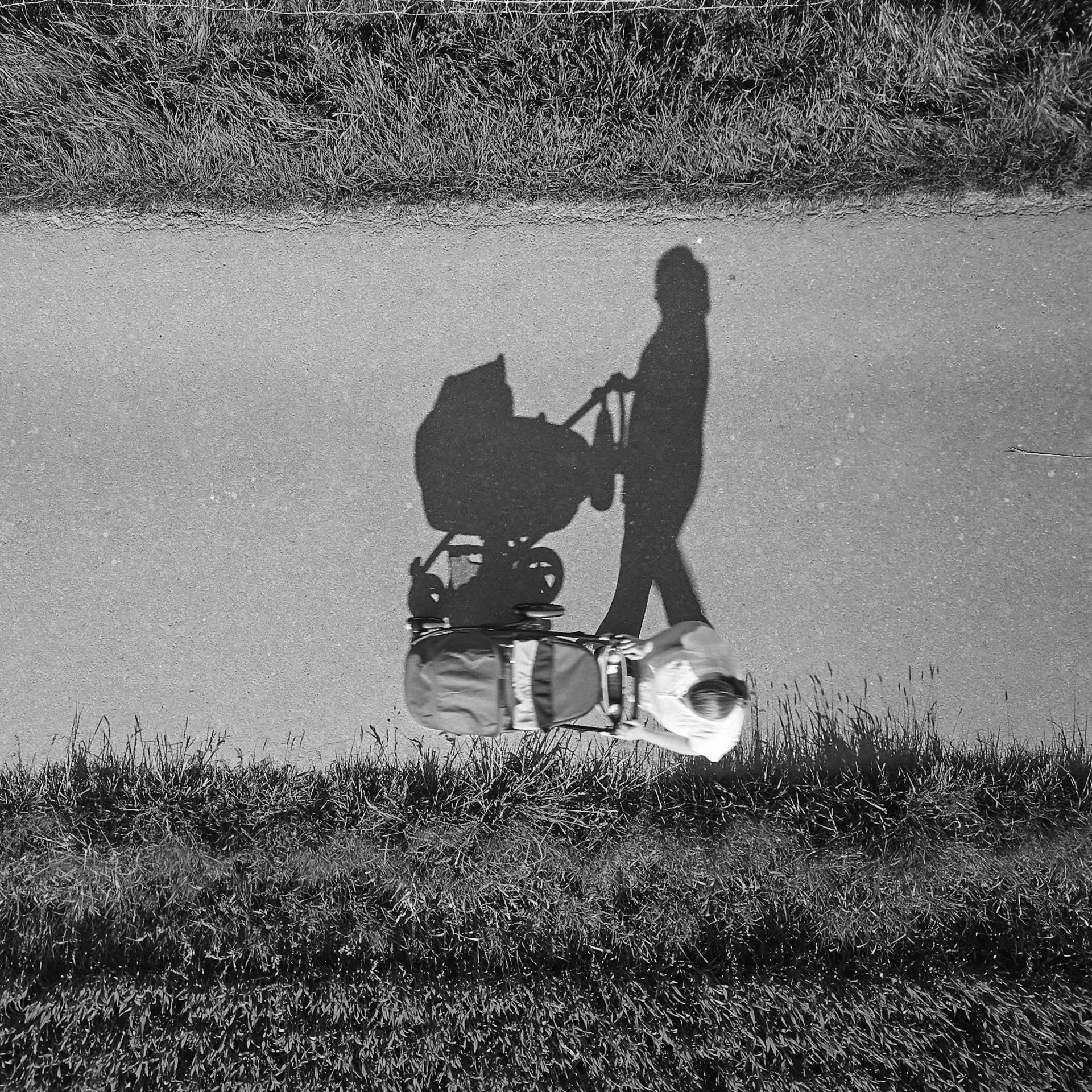 Black and white photograph of a person walking on a paved path, pushing a stroller, with their shadow cast on the ground.