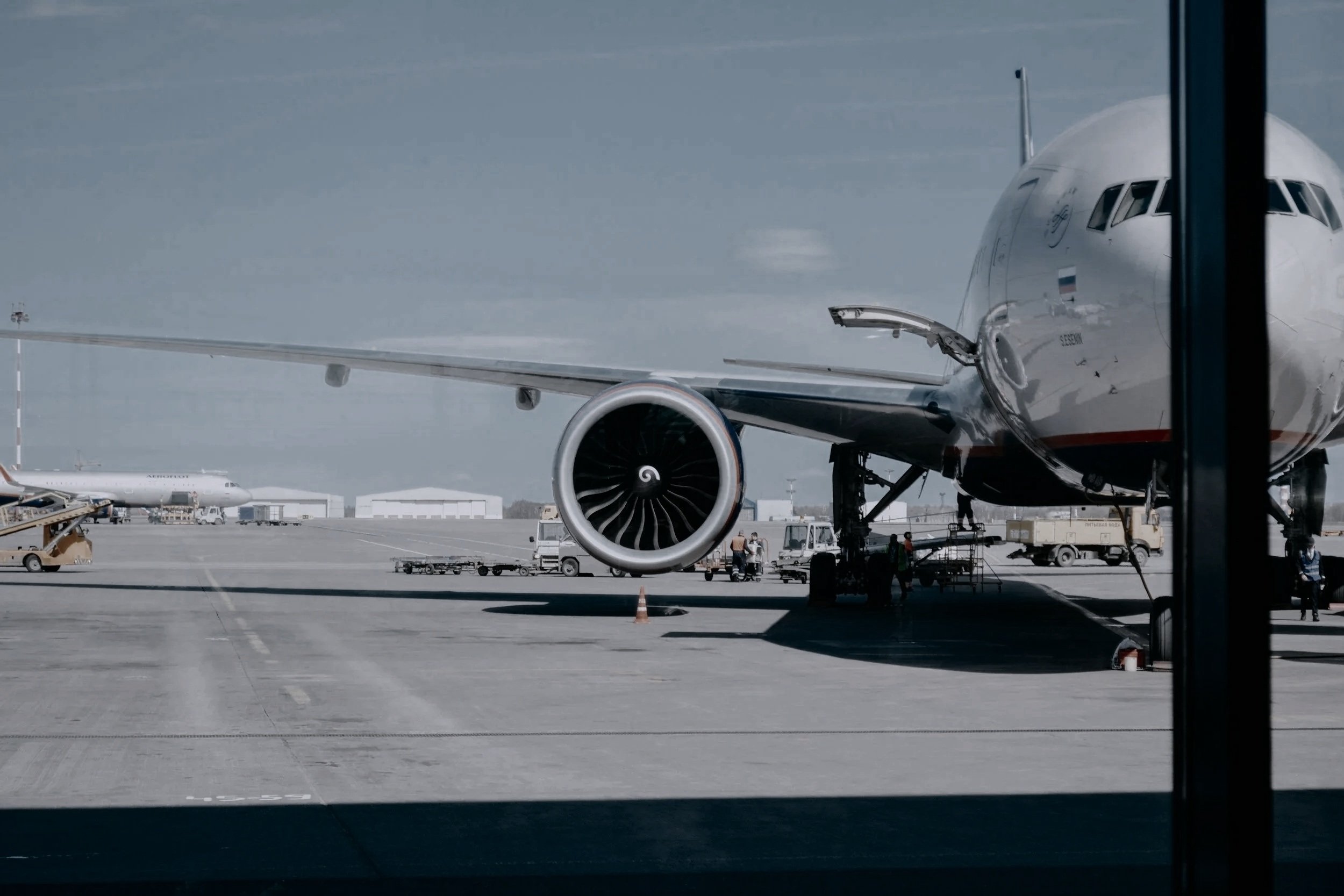 View of an airplane parked at the airport gate from inside the terminal, showing the jet engine and part of the fuselage.