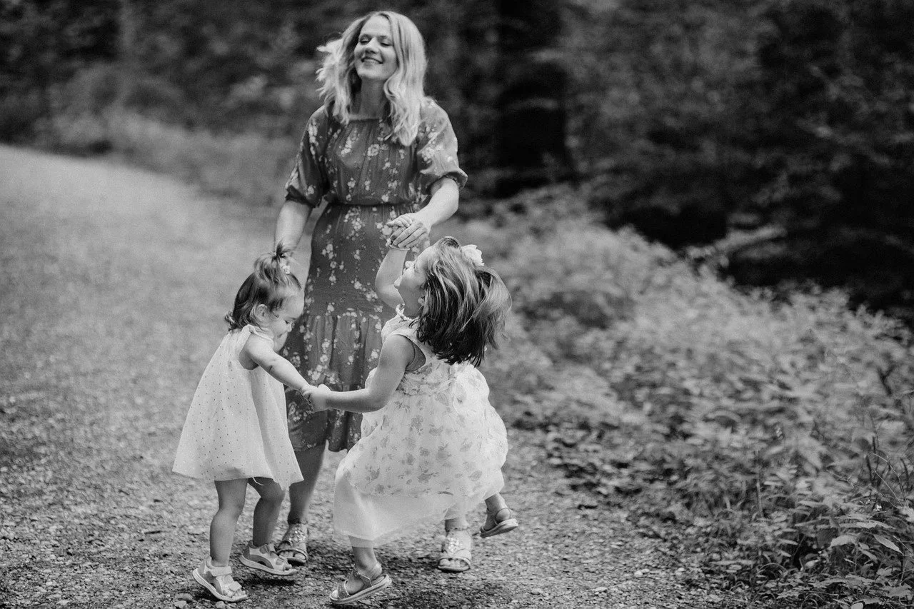 A woman with shoulder-length hair smiling while holding hands with two young girls, dancing and playing on a gravel path in a wooded outdoor setting.