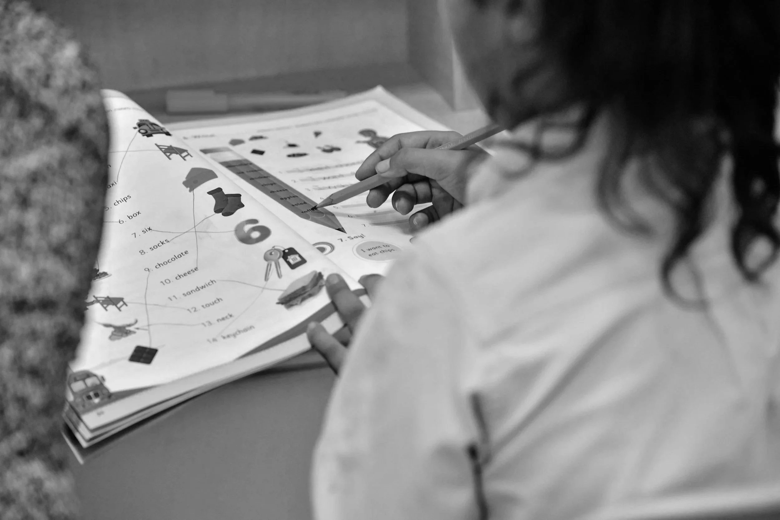 A child working on a colorful educational worksheet with pictures and text, using a pencil, sitting at a table.