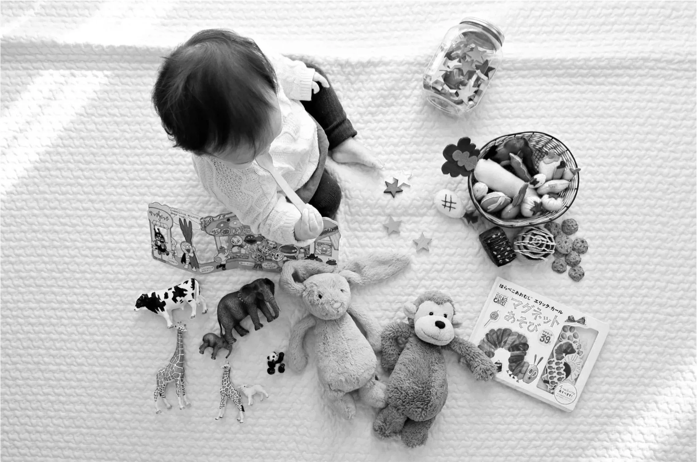 A child sitting on a textured blanket surrounded by toys and books, including stuffed animals, plastic animal figures, a bowl of food items, and a container of star-shaped objects.