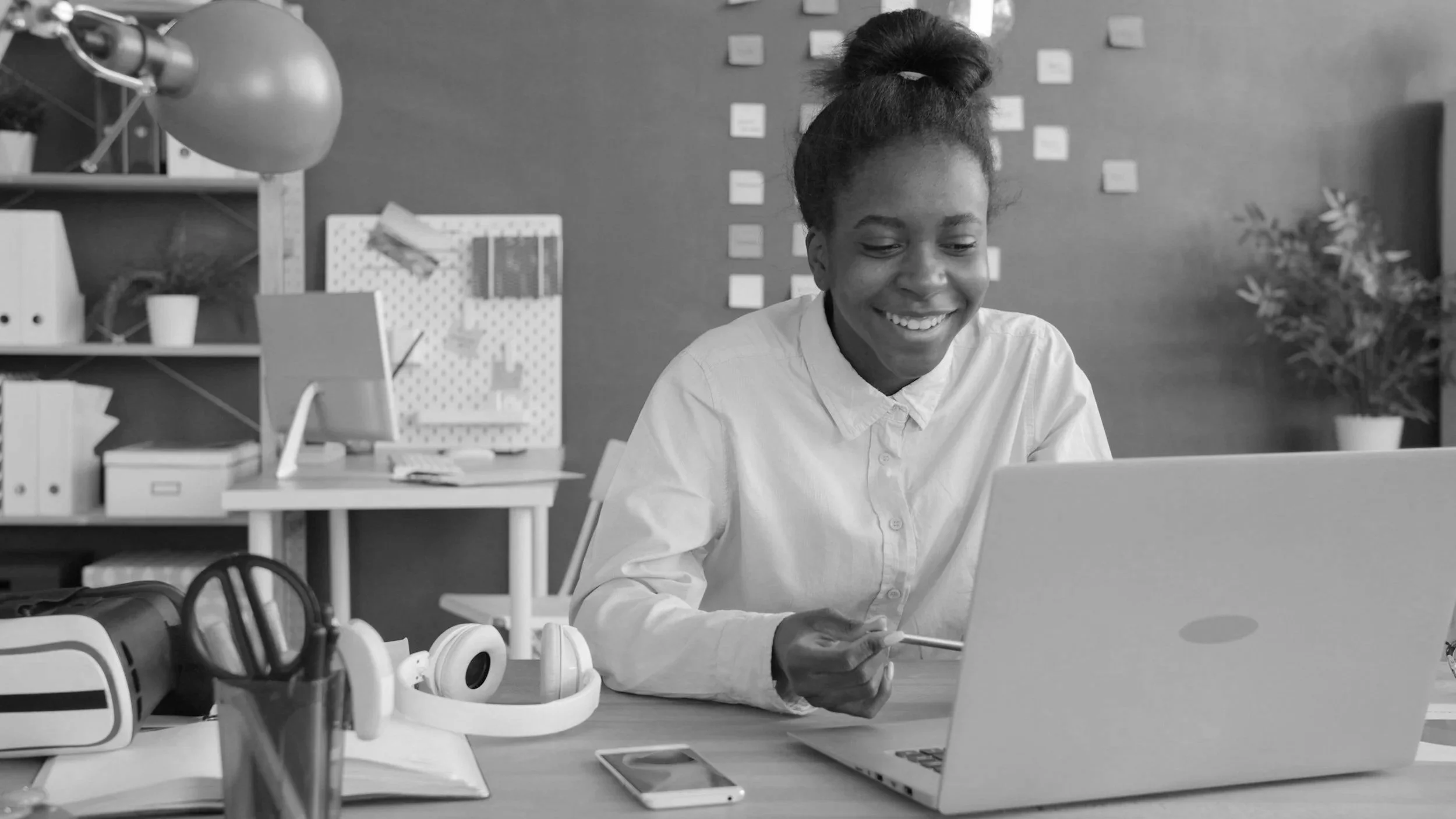 A woman smiling at her laptop in a modern office, with headphones, a smartphone, and office supplies on her desk.