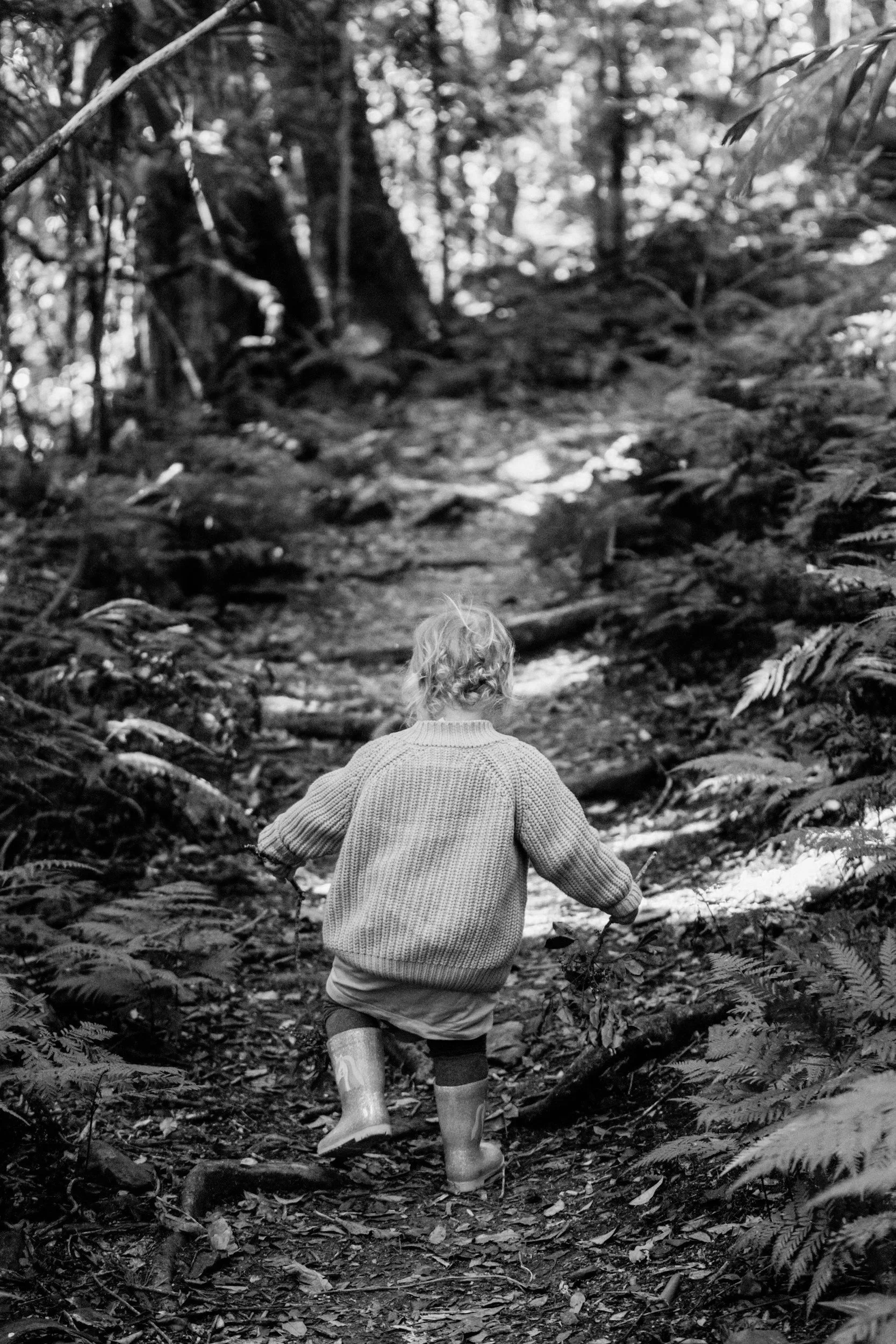 A young child with curly hair wearing a sweater and rain boots walks along a wooded trail in a forest.