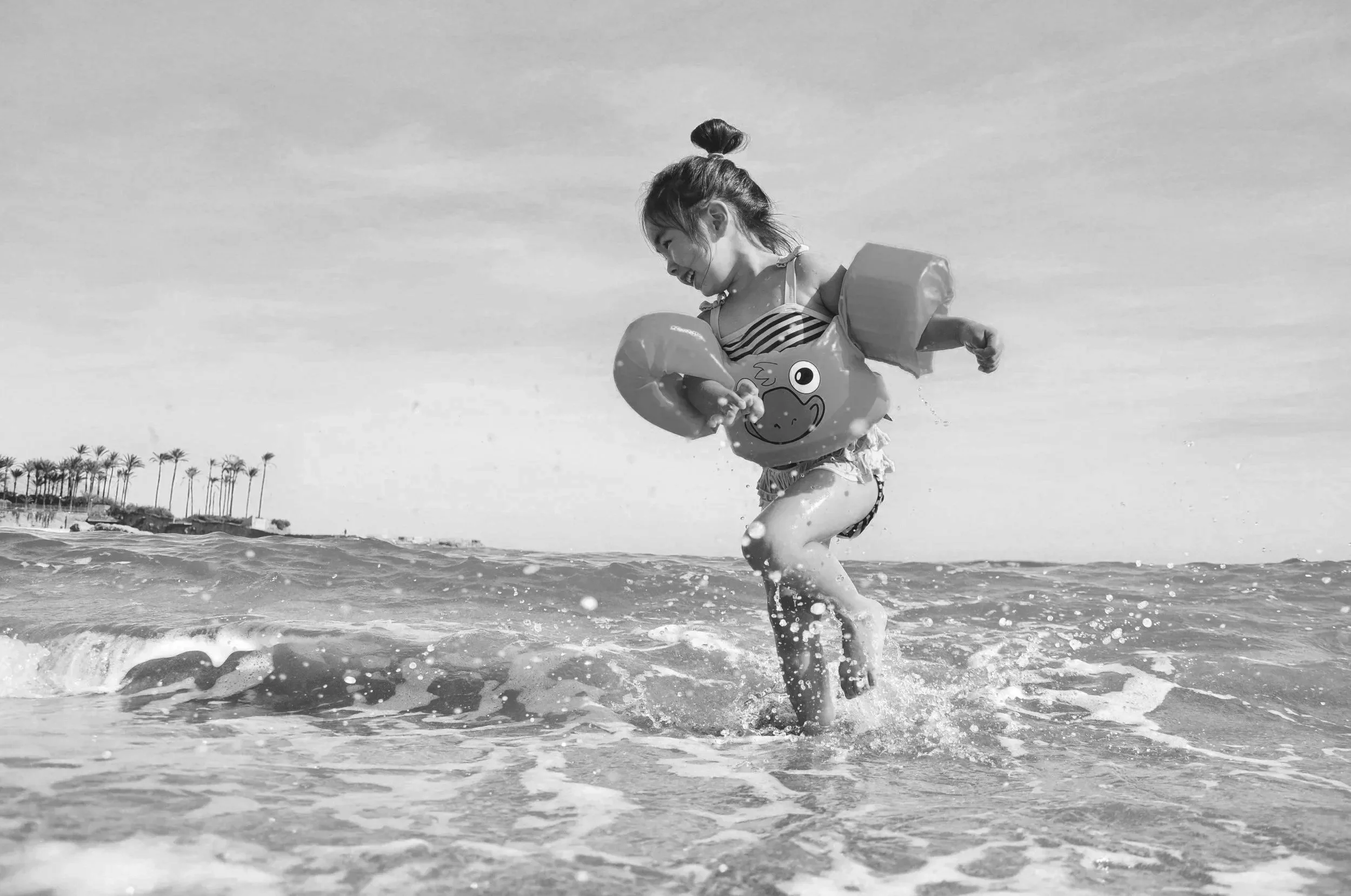 A young girl playing in the ocean water, holding a fish-shaped flotation device, with palm trees visible in the distance on the shoreline.