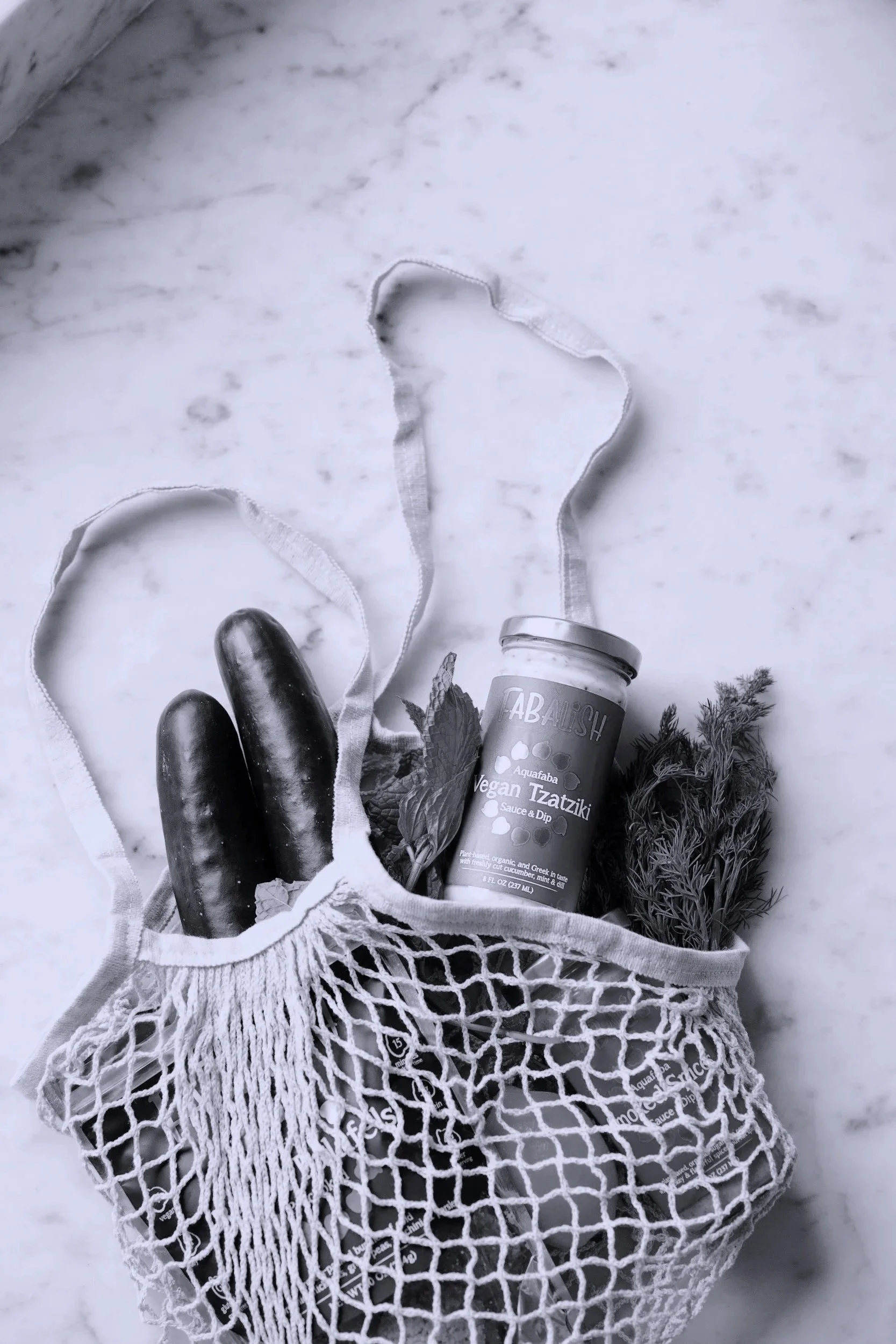 Reusable mesh bag containing two cucumbers, a jar of vegan tzatziki sauce, and some fresh herbs, resting on a marble surface.