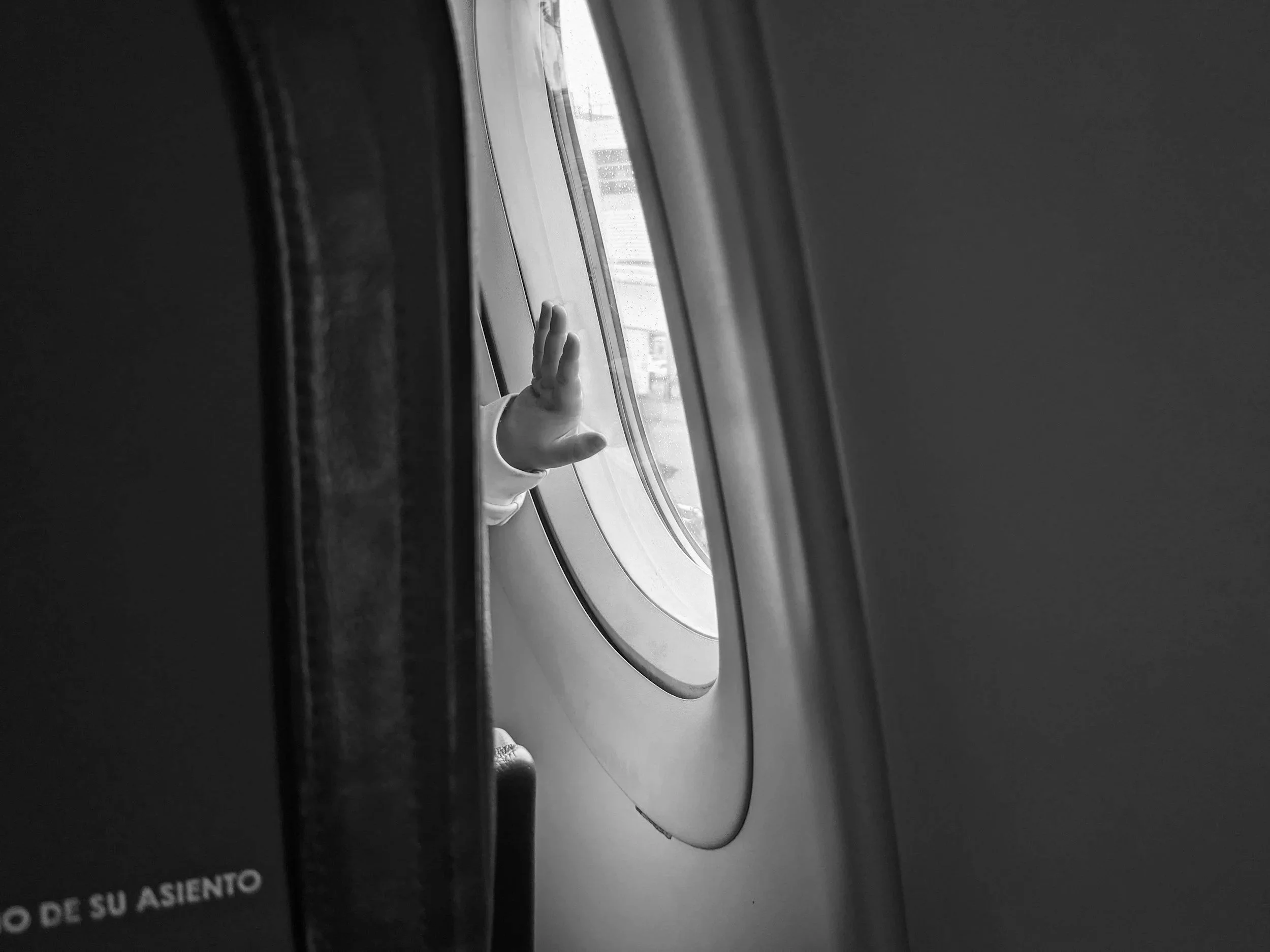 A person’s arm and hand with a white sleeve reaching out of an airplane window, making a peace sign with their fingers.