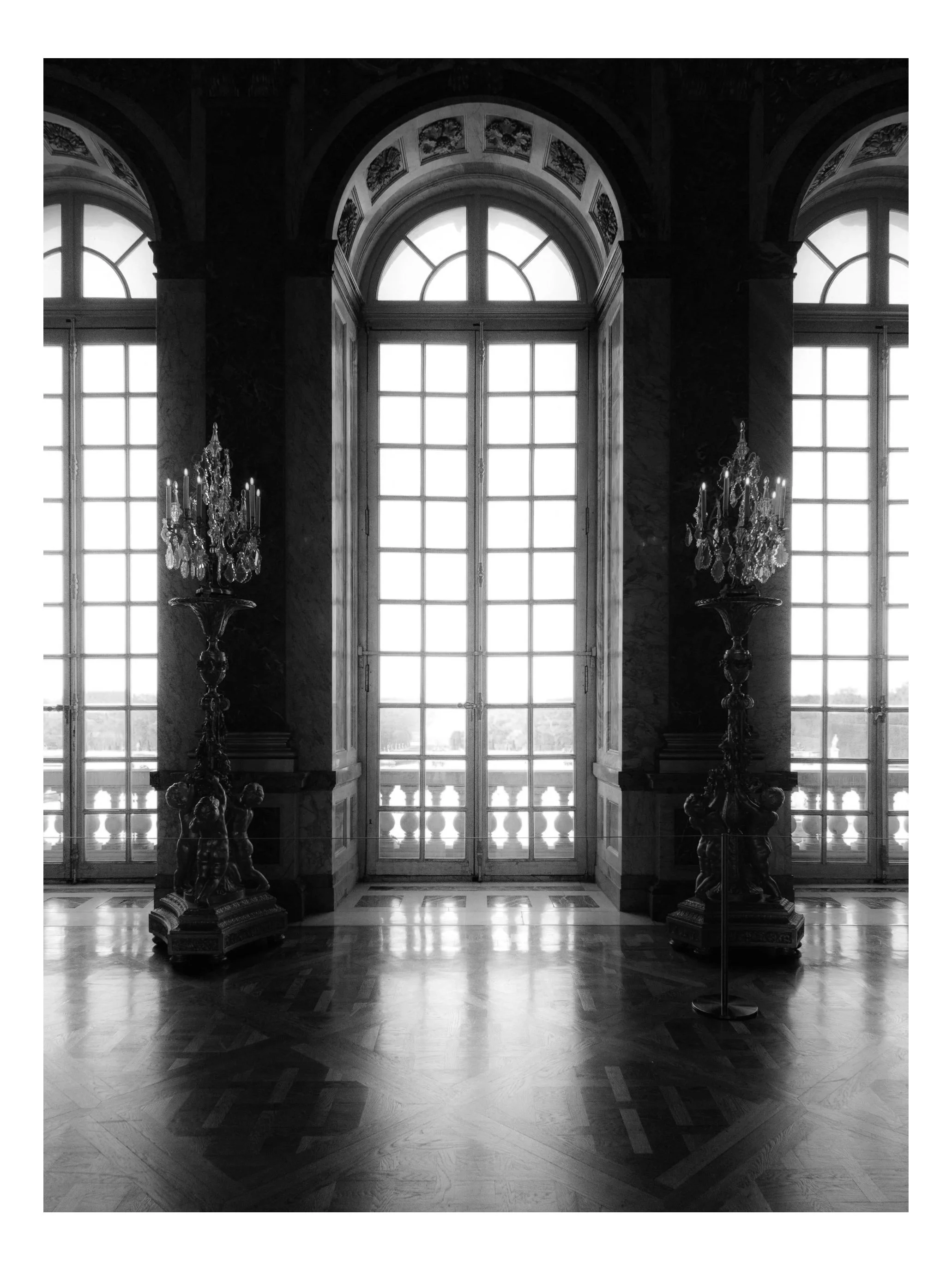 Interior of a grand room with tall arched windows, ornate chandeliers with crystal decorations, dark marble walls, and an intricately patterned wooden floor, with a view outside through the windows.