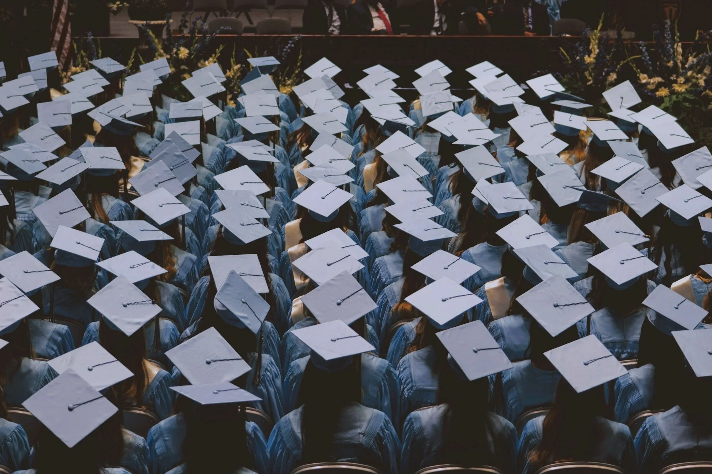 A large graduation ceremony with students wearing caps and gowns seated in rows facing a stage, with floral arrangements in the background.