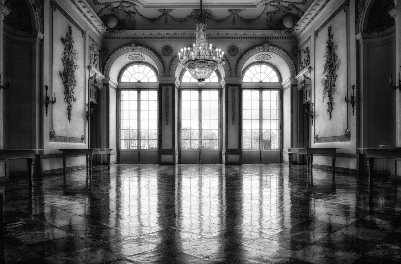 An elegant, historic ballroom with large windows, ornate ceiling decorations, a crystal chandelier, and a reflective wooden floor.