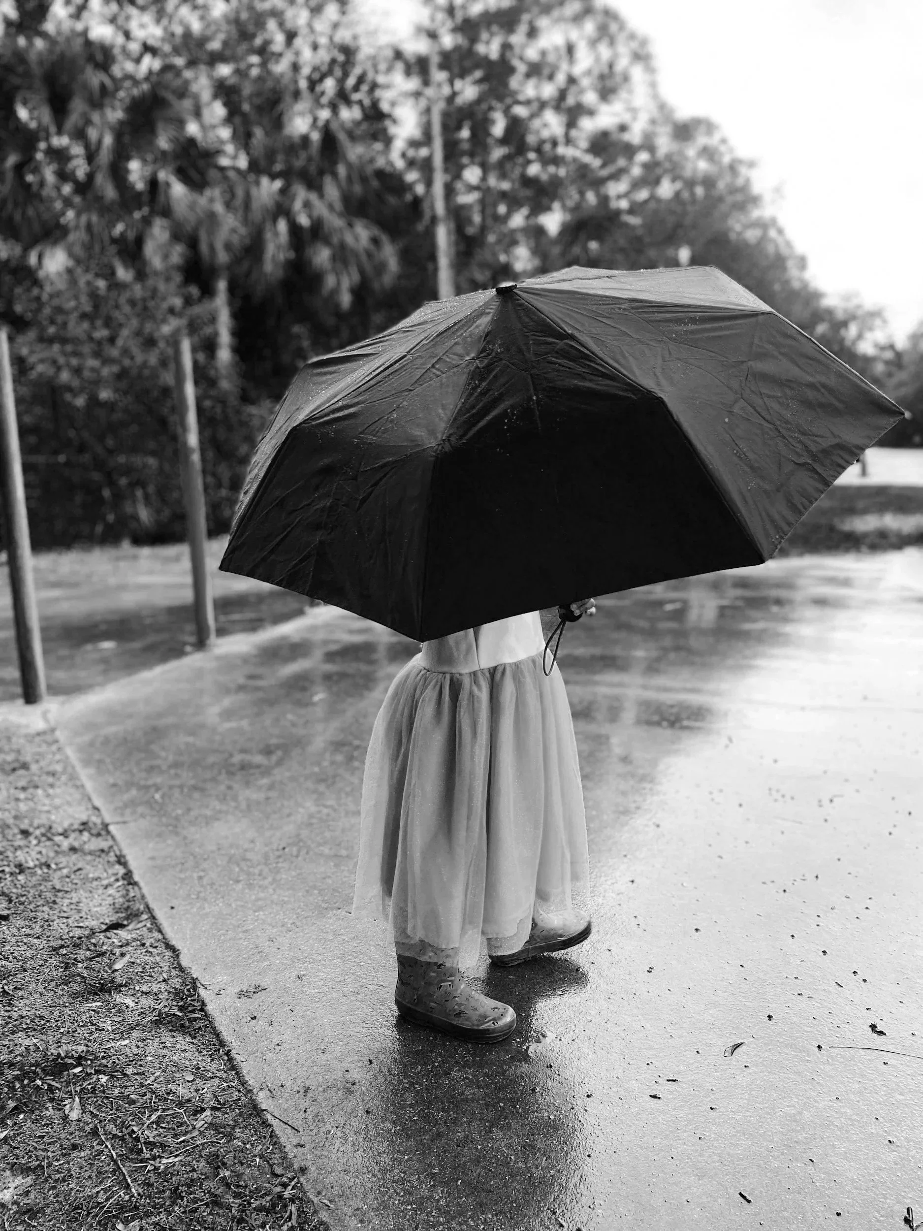 A young girl holding a large umbrella on a wet sidewalk during rainy weather, wearing rain boots and a light-colored skirt.
