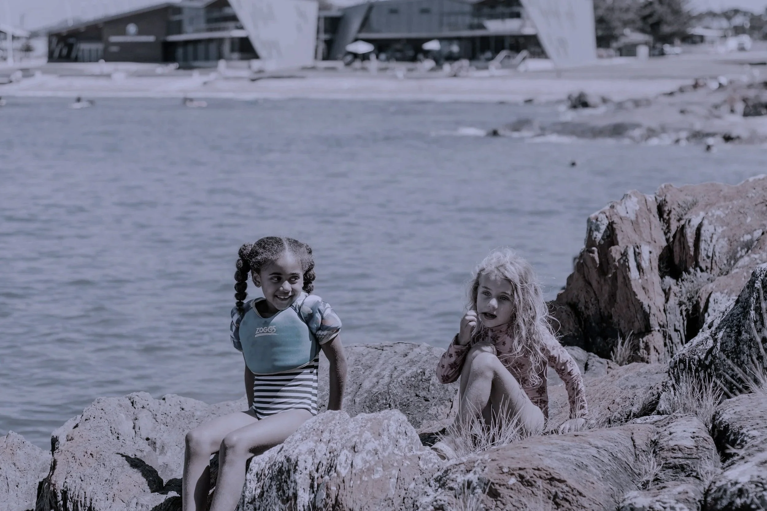 Two young girls sitting on rocks by a body of water, with a modern building in the background.