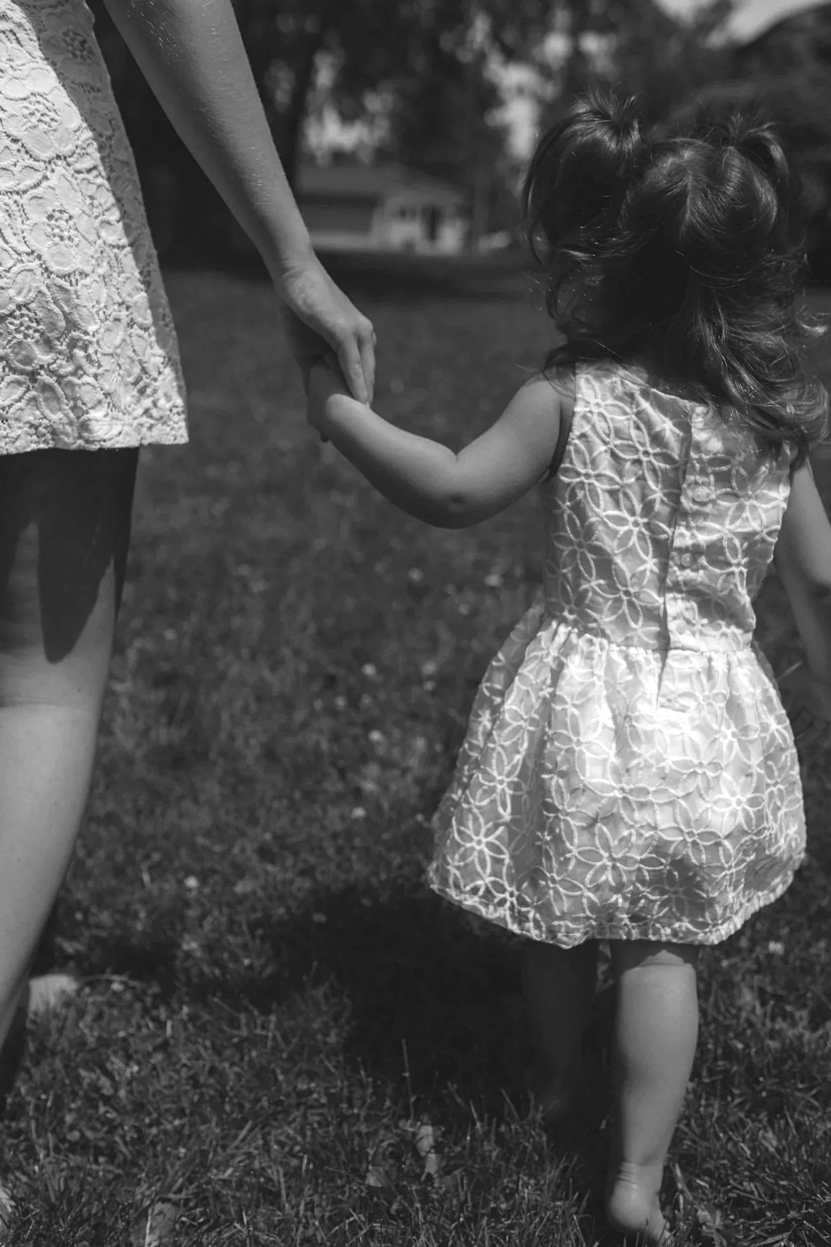 A young girl holding hands with an adult, walking outdoors on a grassy area on a sunny day, both wearing dresses with lace patterns.
